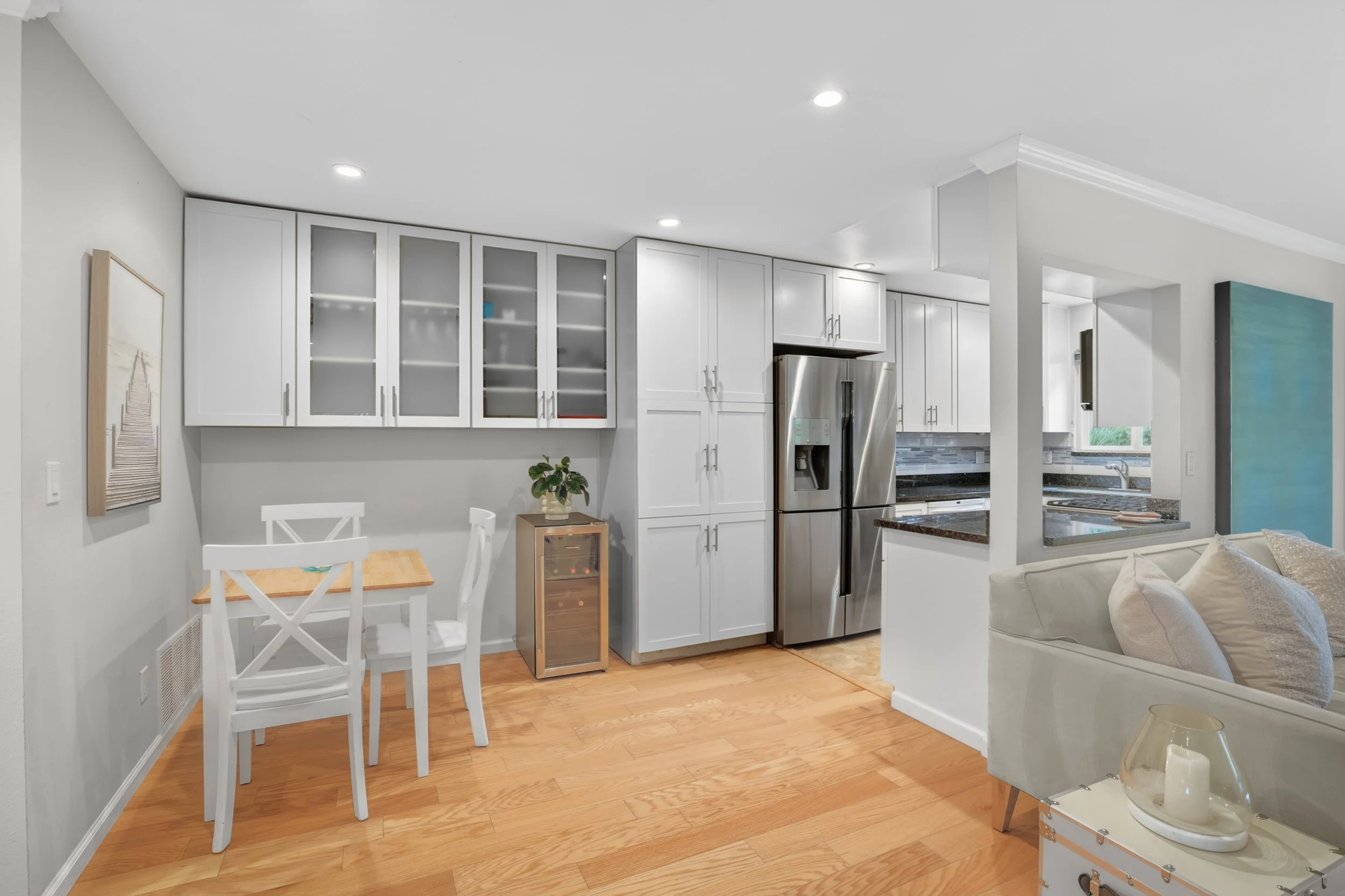 Modern kitchen with white cabinets, stainless steel refrigerator, black granite countertops, small dining table with three chairs, wooden flooring, and a living area with a white sofa and a glass lantern