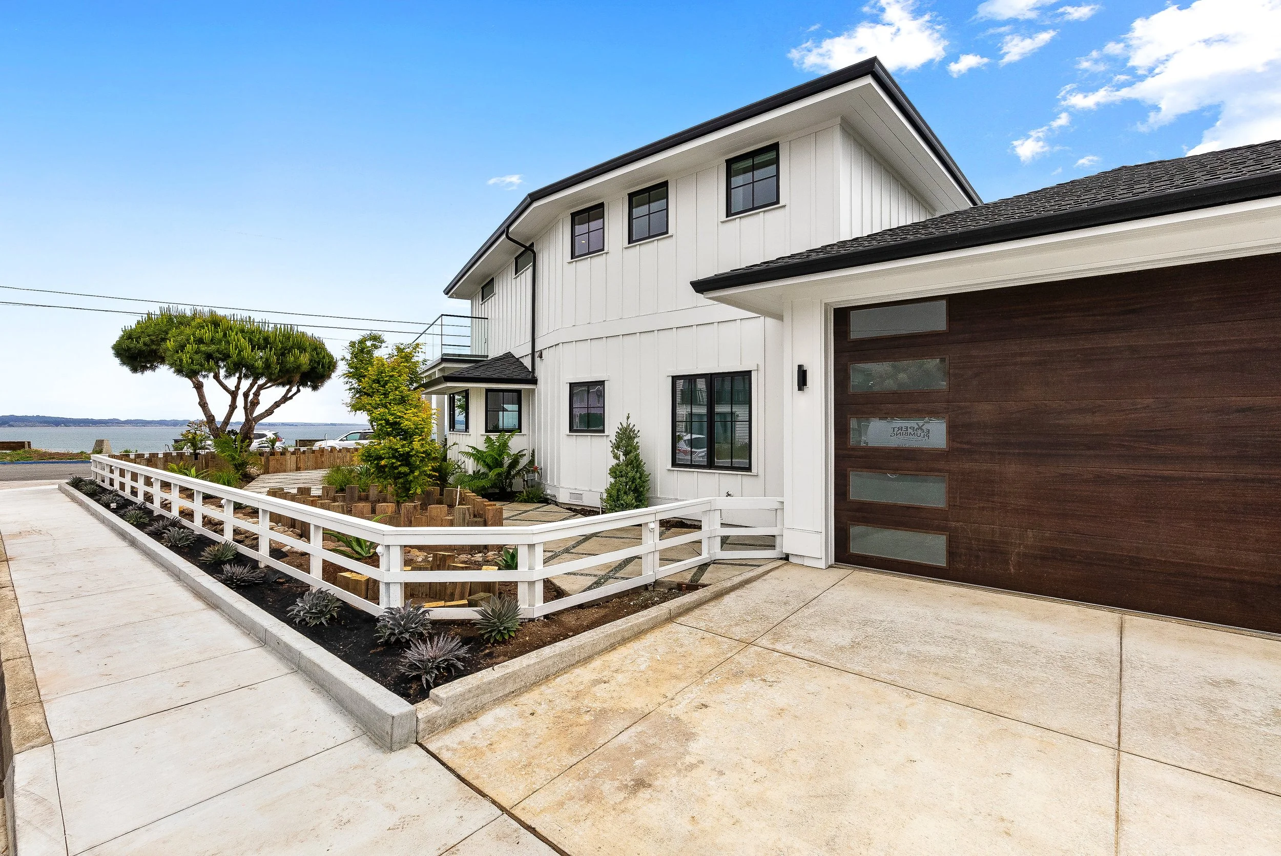 Modern two-story house with white vertical siding, dark wood garage door, and landscaped front yard with trees, plants, and a white fence, near a body of water under a blue sky in Capitola.