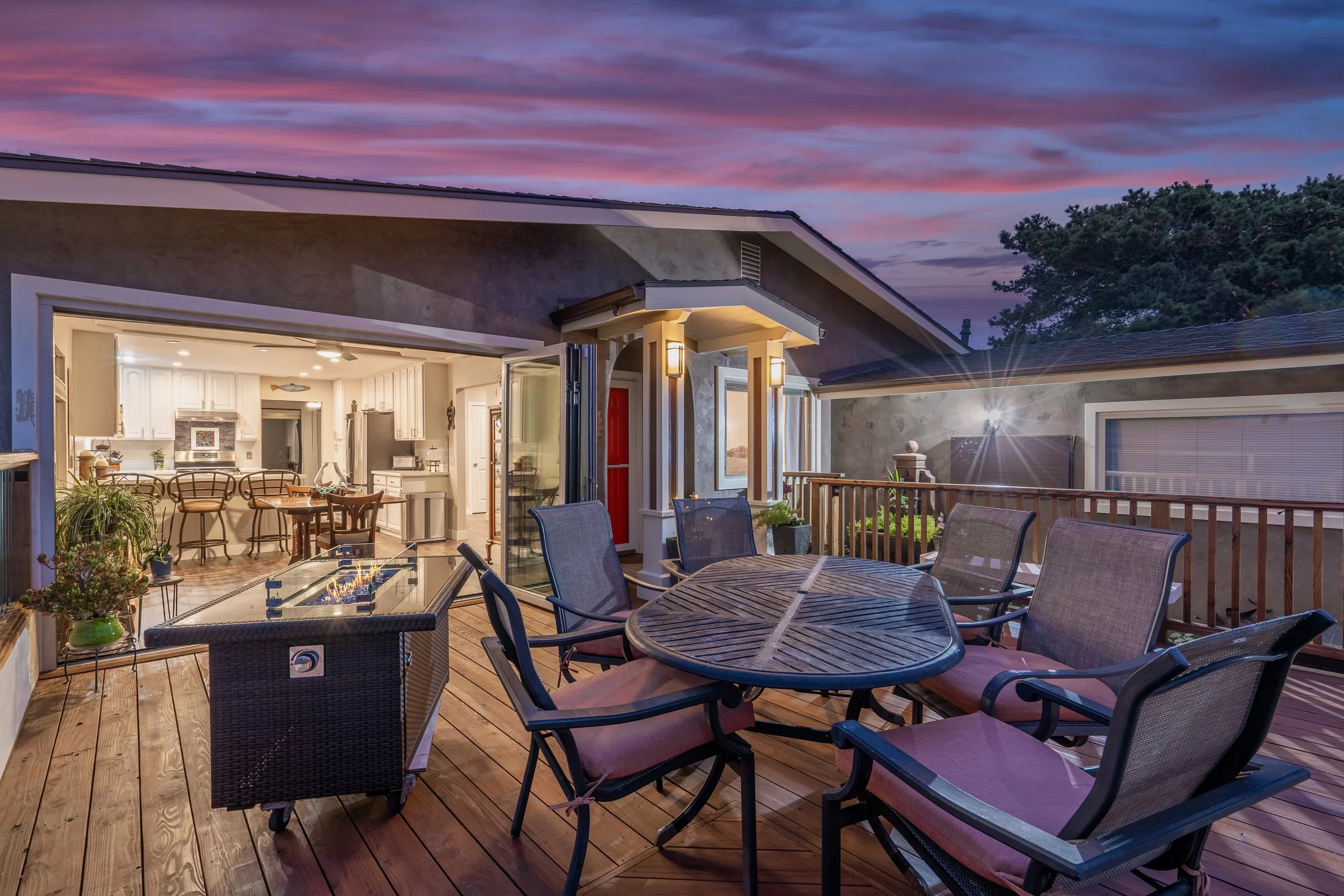 Outdoor wooden deck with a round dining table and six chairs, fire pit table, and potted plants, leading into a well-lit kitchen and living area of a house at dusk, with a colorful sky.