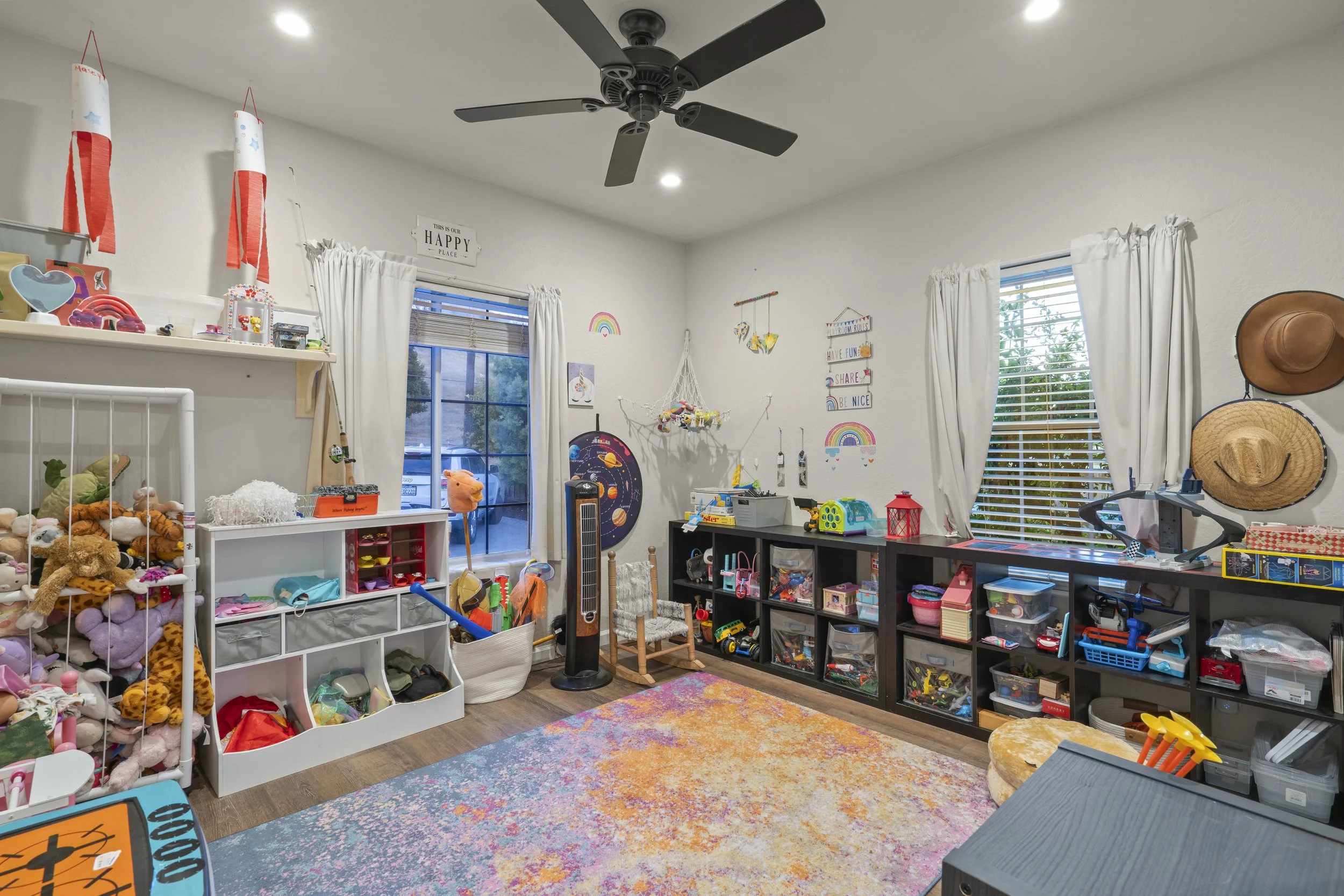 A cluttered child's playroom with toys, stuffed animals, shelves, windows with white curtains, and colorful wall decorations.