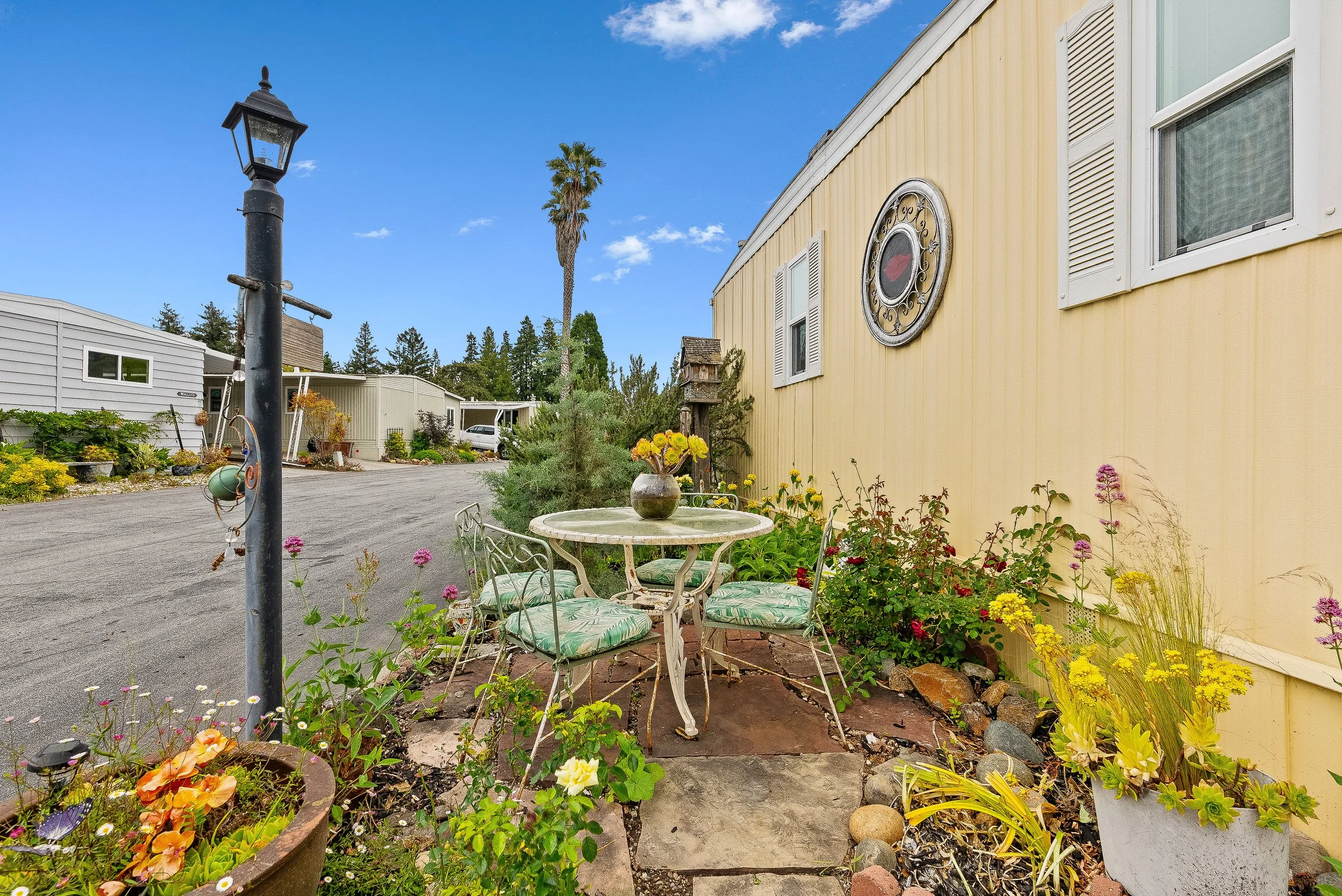 Yard with outdoor metal table and chairs, potted flowers, plants, and a yellow mobile home, under a blue sky with clouds.