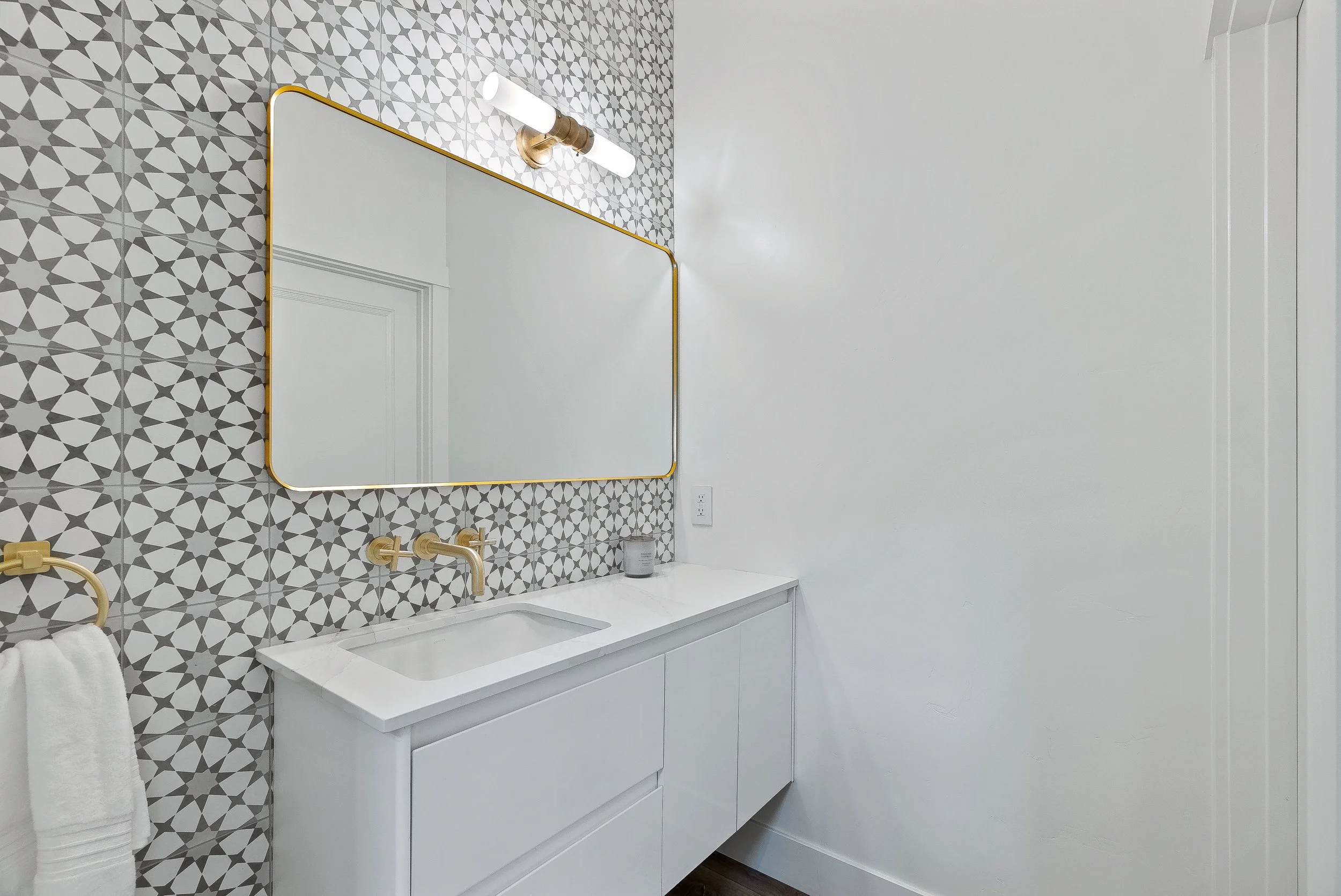 Modern bathroom with patterned tile wall, large mirror, white vanity, gold fixtures, and a towel on a rack in Capitola, California.