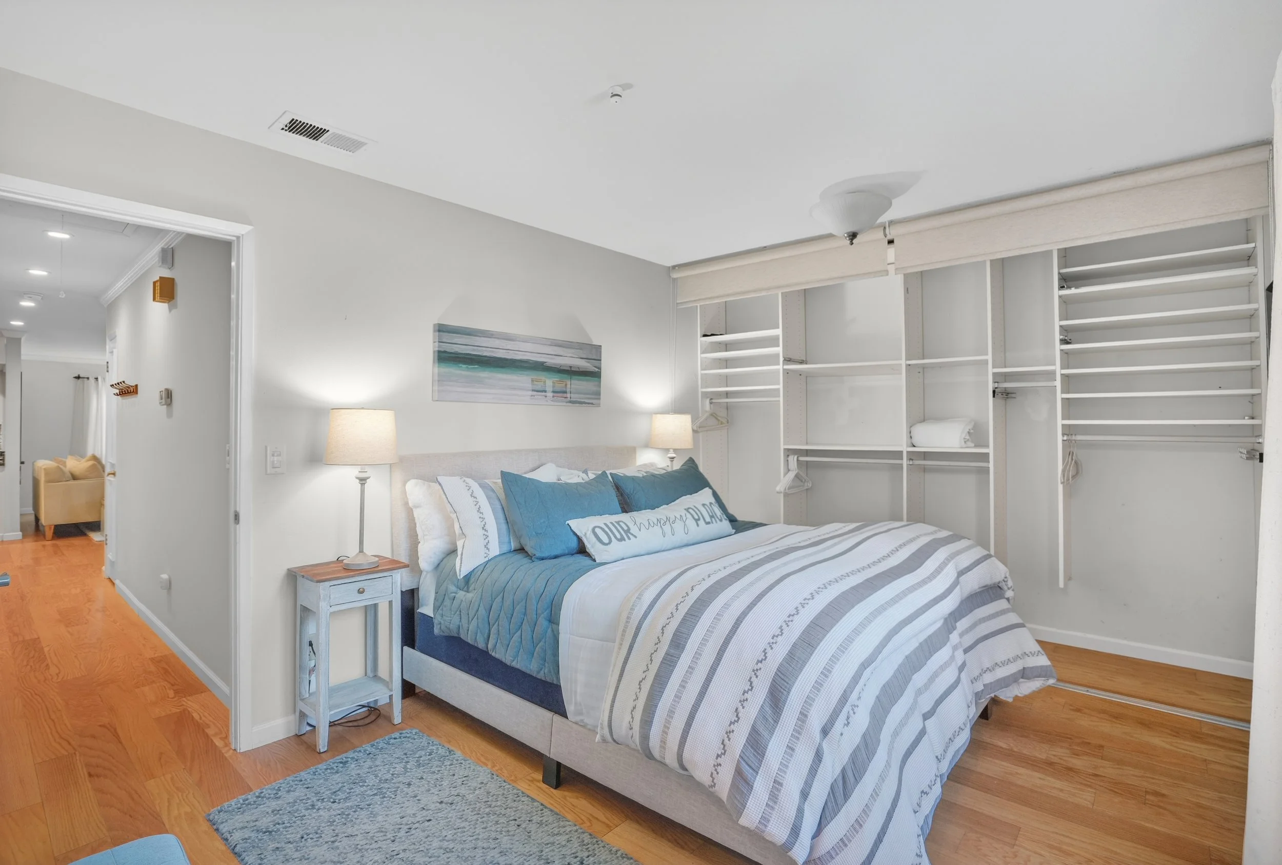 A bedroom with a bed featuring a blue and white striped comforter, blue pillows, and a decorative pillow reading 'Our Happy Place.' There is a bedside table with a lamp on each side of the bed, and an open closet with empty shelves and some hanging s