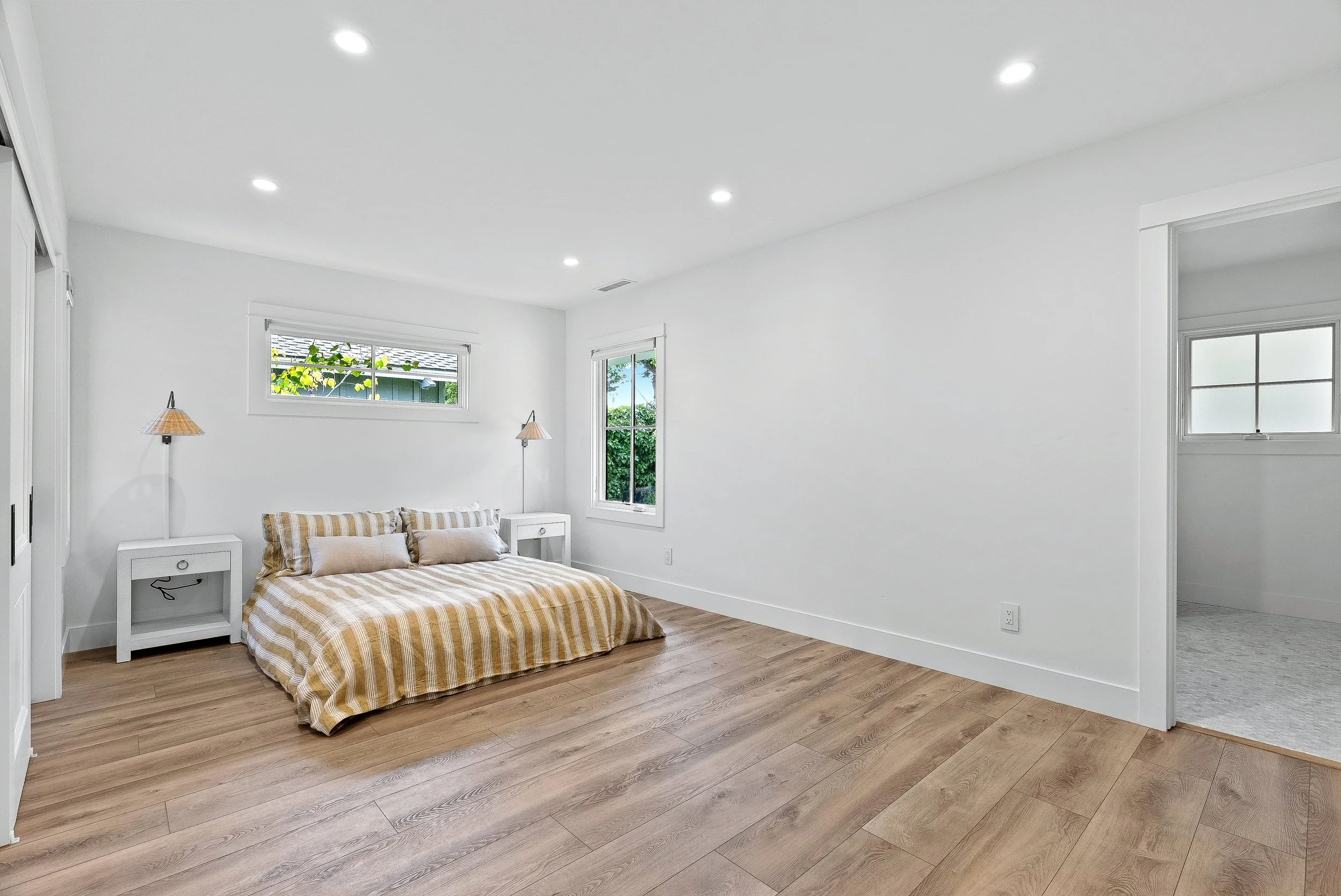 A minimalist bedroom with white walls, wooden floor, a bed with striped bedspread, two side tables with lamps, and windows showing green outdoors in Capitola, California.