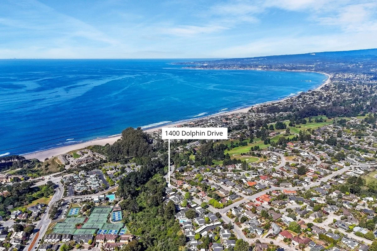 Aerial view of a coastal neighborhood with houses, trees, and tennis courts near the ocean shoreline with a label pointing to 1400 Dolphin Drive.
