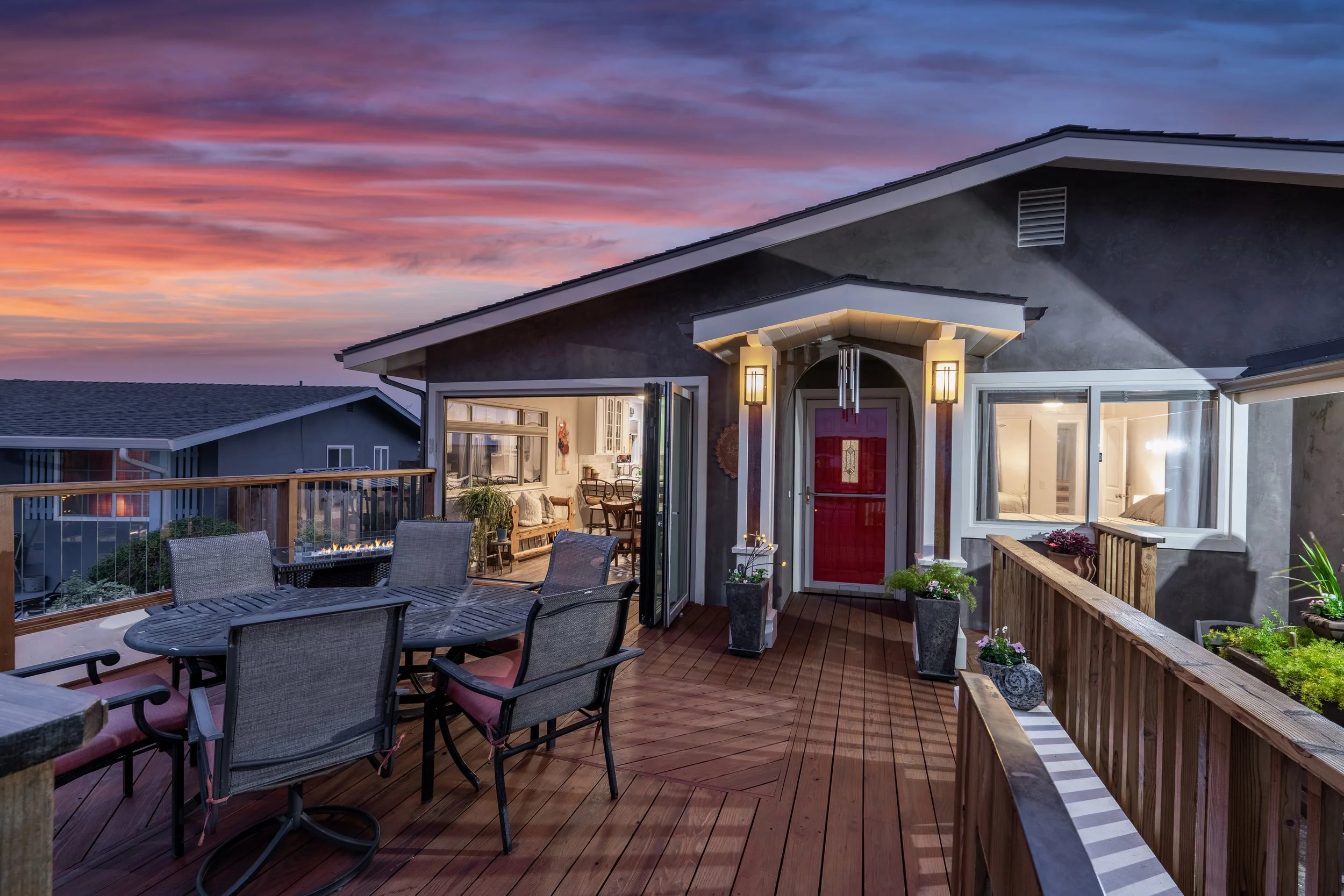 A cozy outdoor wooden deck at dusk with a round table and chairs, potted plants, and a glowing fire feature, attached to a house with a view into a well-lit living room and bedroom.