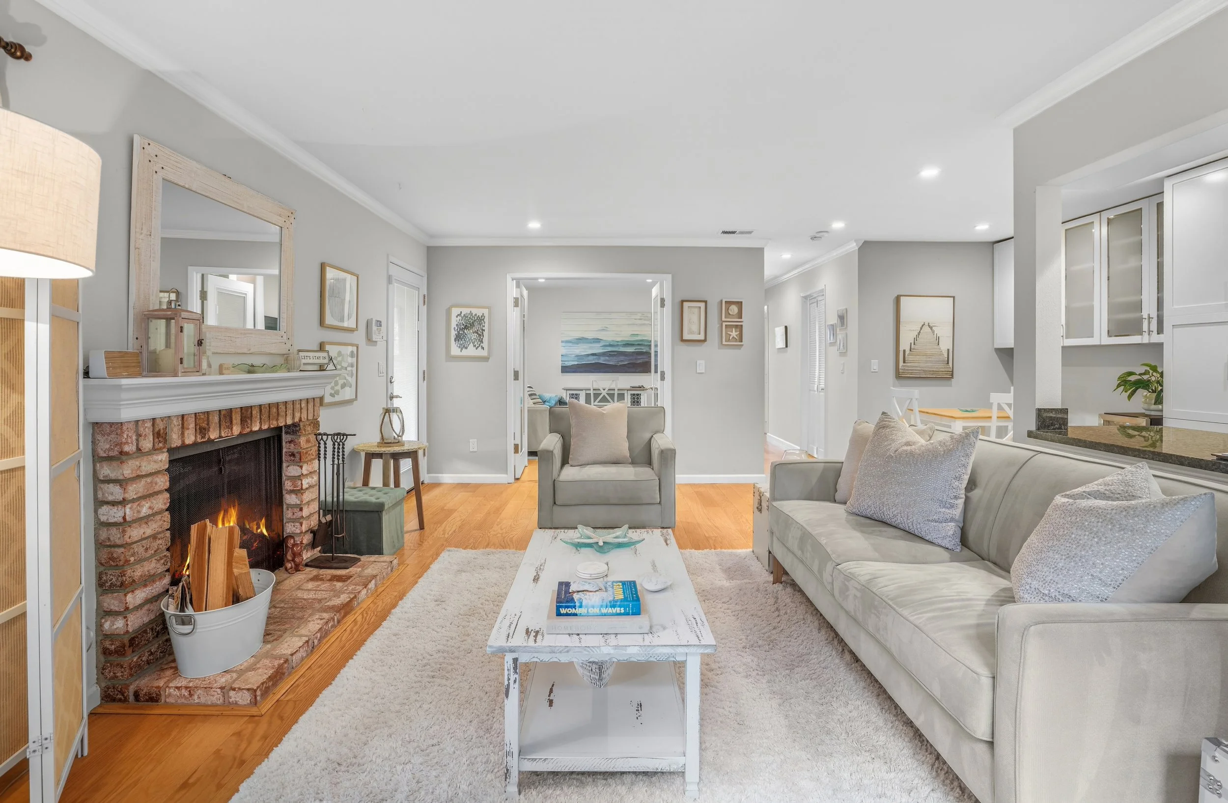Living room with fireplace, beige sofa, armchair, white coffee table with books and decor, wooden flooring, light gray walls, artwork, and a view into another room with a dining area.