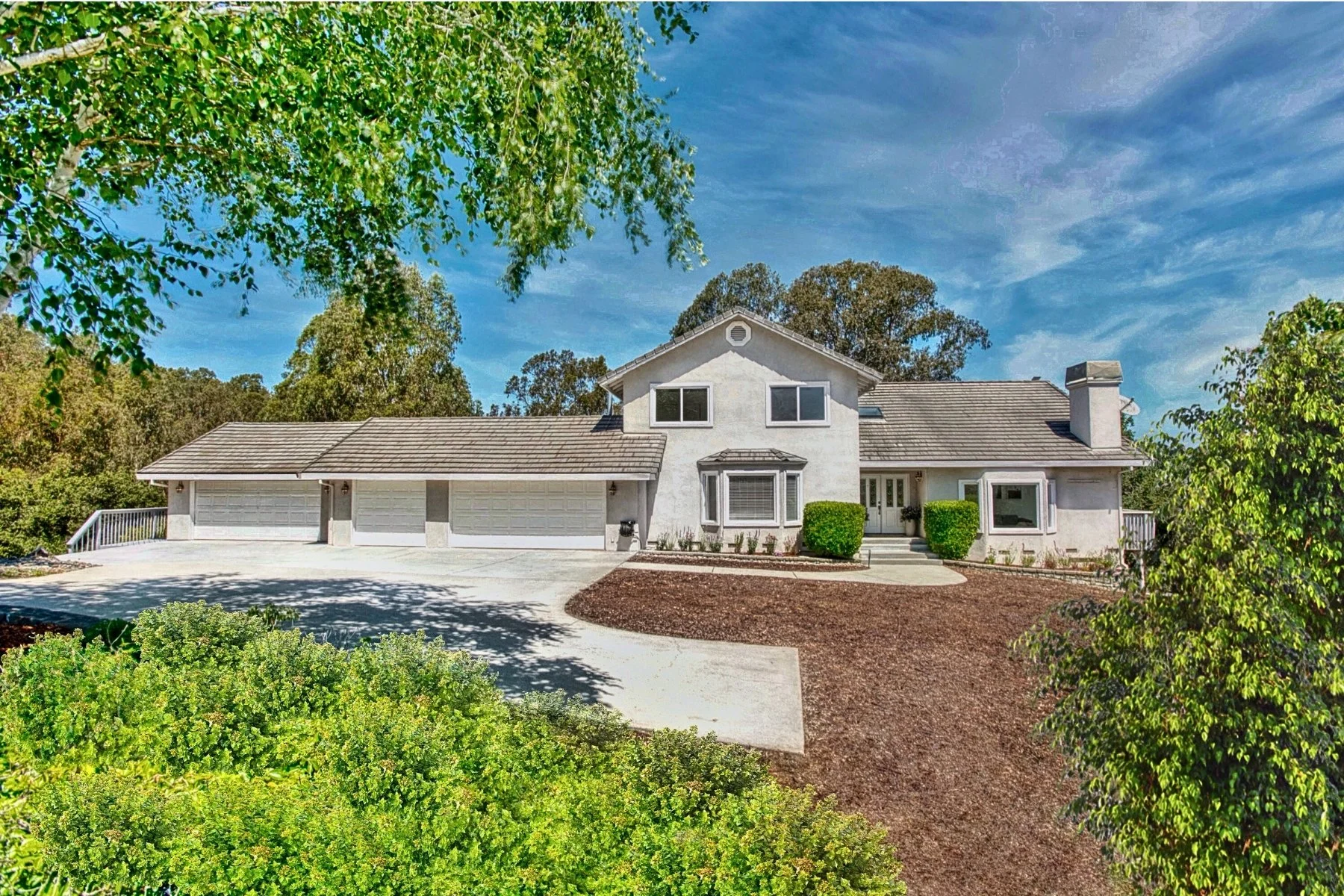 Large white house with attached garage, surrounded by green bushes and trees, under a blue sky with some clouds.