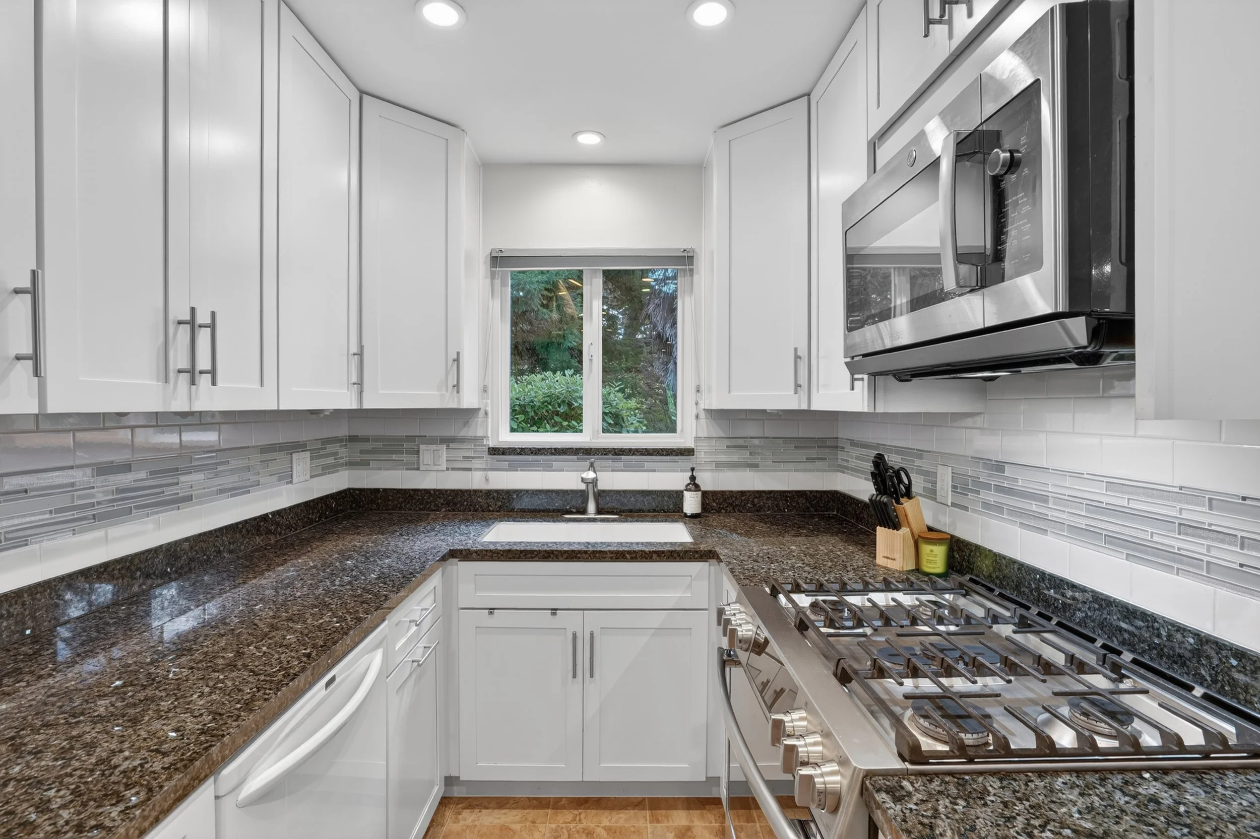 Kitchen with white cabinets, brown granite countertops, stainless steel microwave and stove, white tile backsplash, window above sink, and some utensils and soap on the counter.