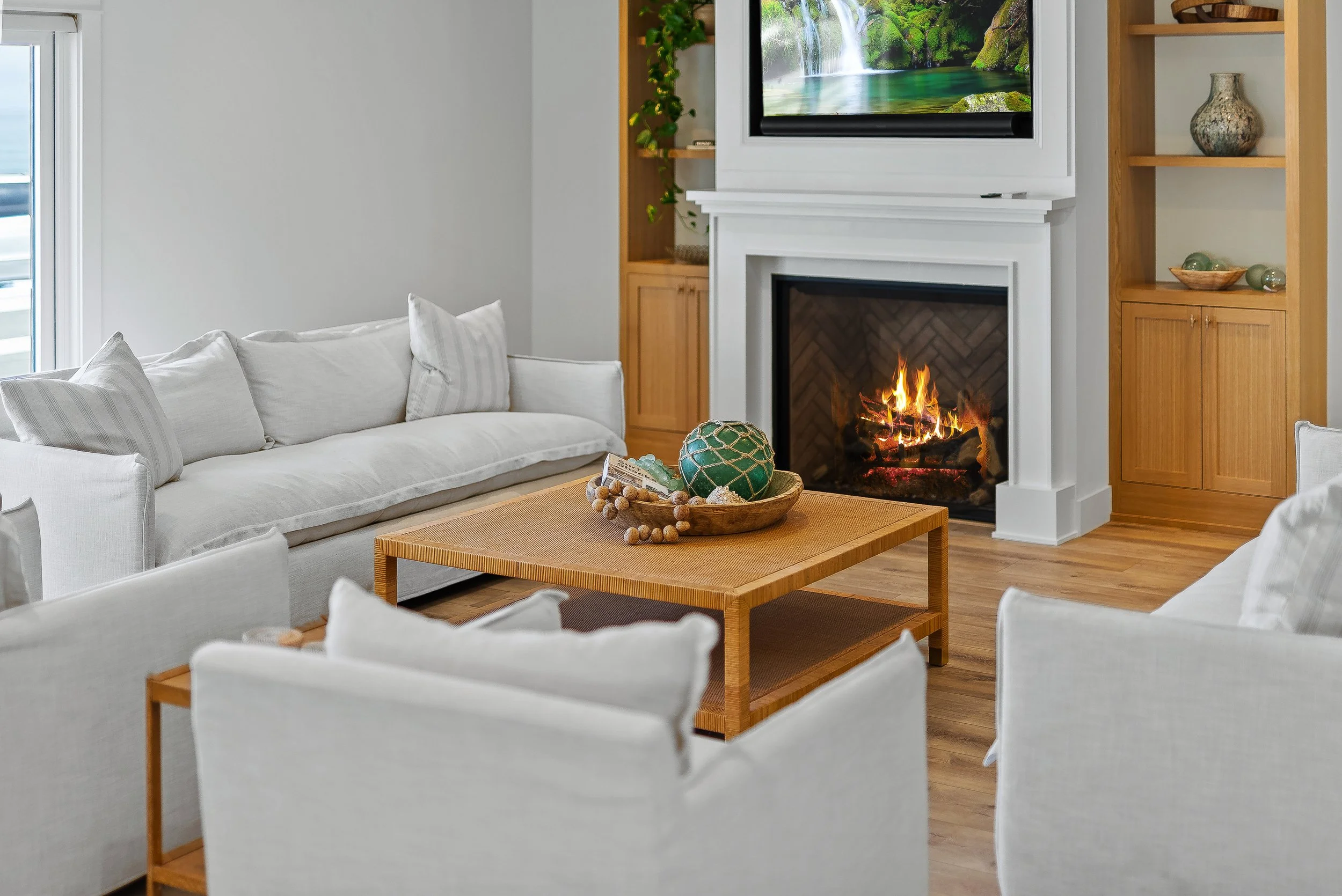 Living room with white sofas, a wooden coffee table, a fireplace with a fire, a wall-mounted TV displaying a nature scene, and built-in wooden shelves with decorative items in Capitola, California.