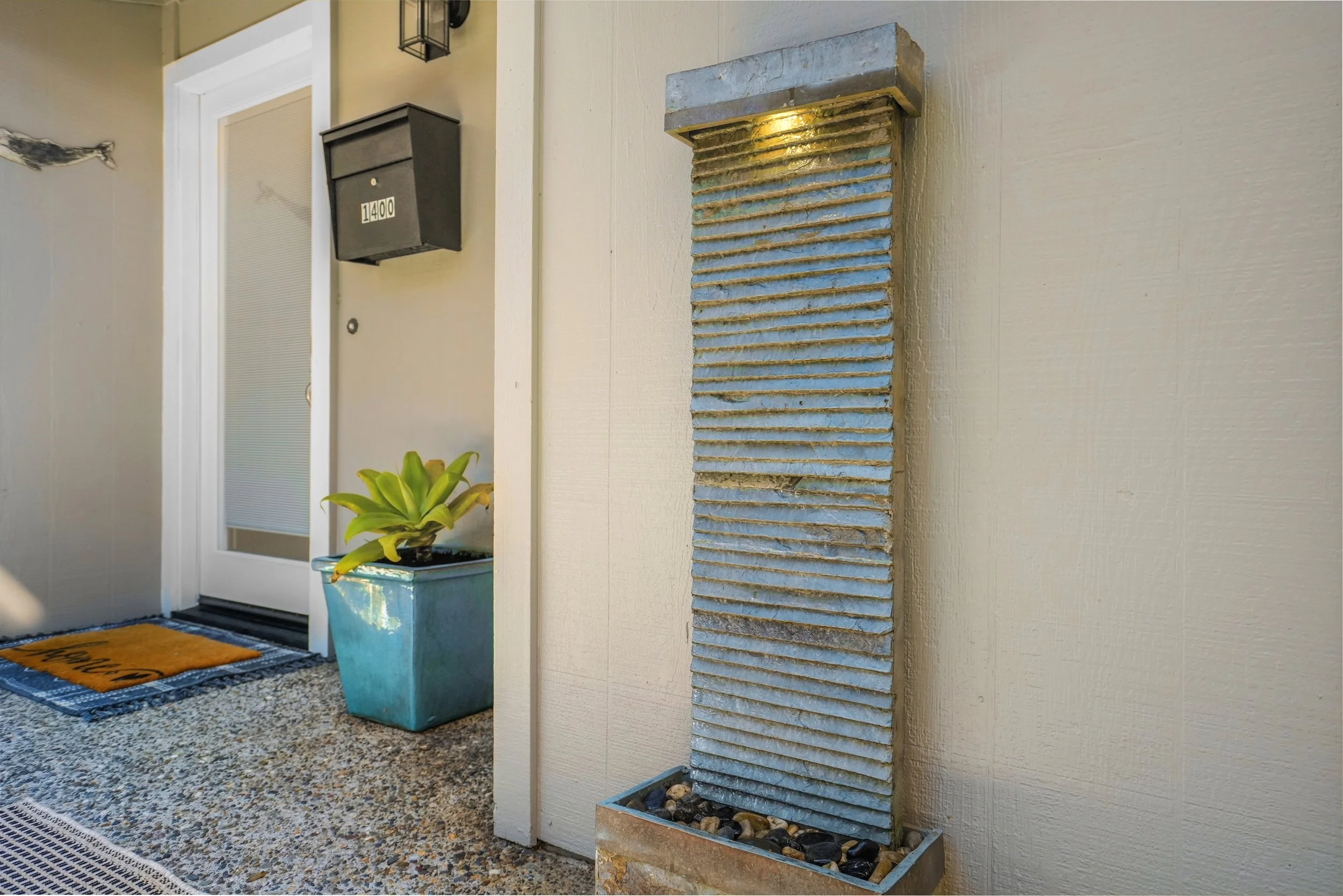Decorative outdoor water feature with a vertical panel of textured, blue-gray stone, water flowing down from the top, placed in a rectangular container filled with small stones, on a porch or patio.