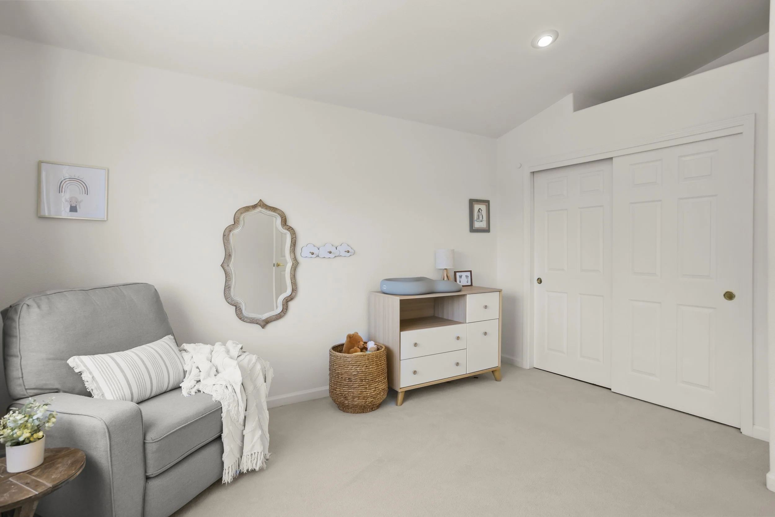 A cozy room with a gray armchair draped with a white blanket and striped pillow, side table with a plant, a woven basket with stuffed toys, a white dresser with a changing pad on top, small framed pictures, wall art, a decorative mirror, and closed c