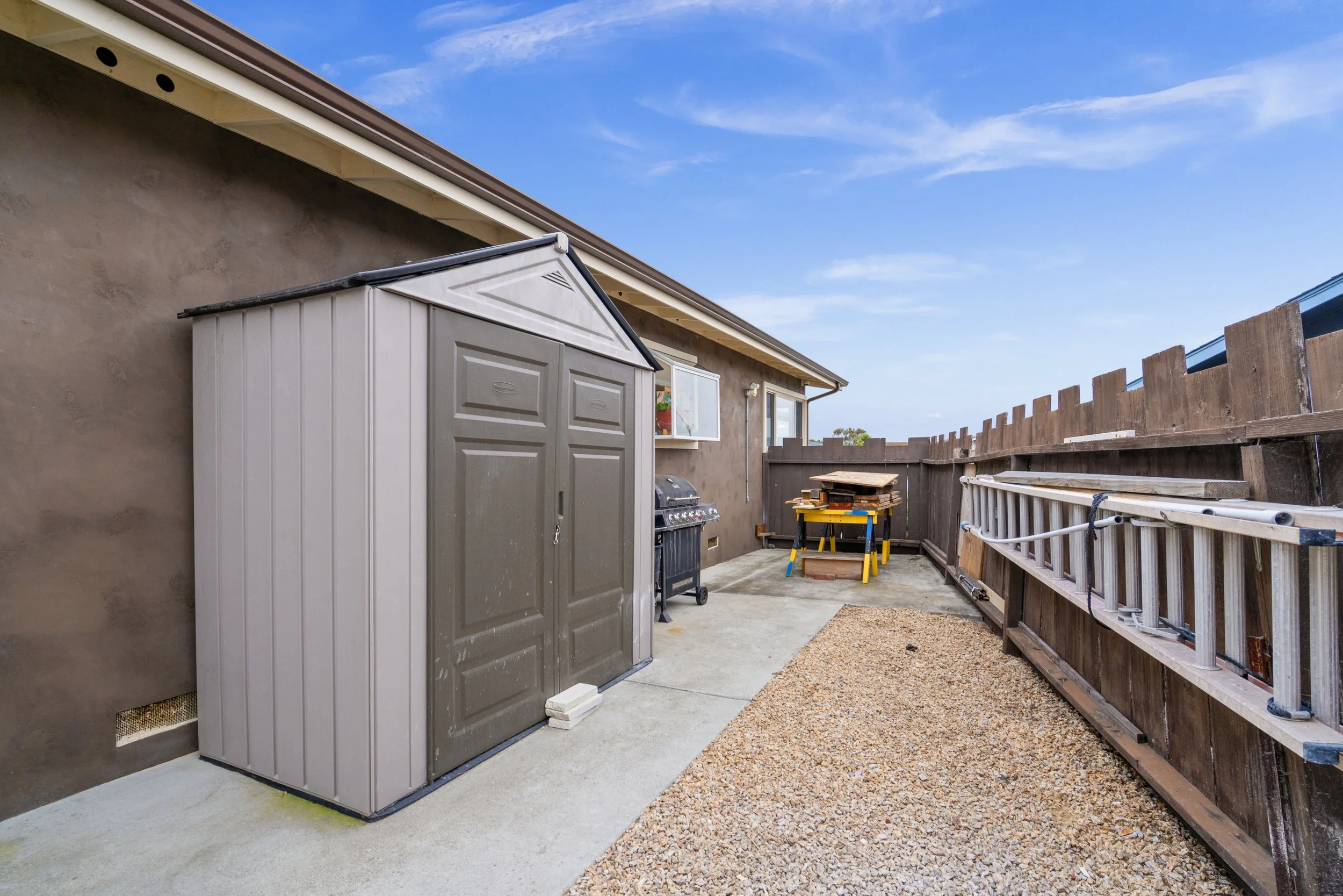 Backyard patio with a storage shed, a barbecue grill, a stack of wooden pallets, a ladder, and a wooden fence under a blue sky.