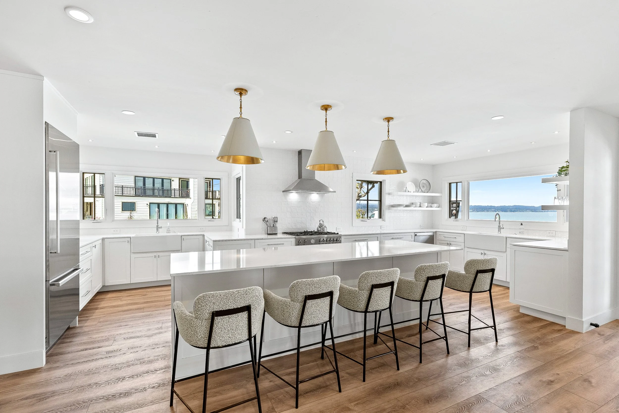 Bright white kitchen with large island, four brunch chairs, stainless steel refrigerator, modern stove, and large windows overlooking water Capitola, California.