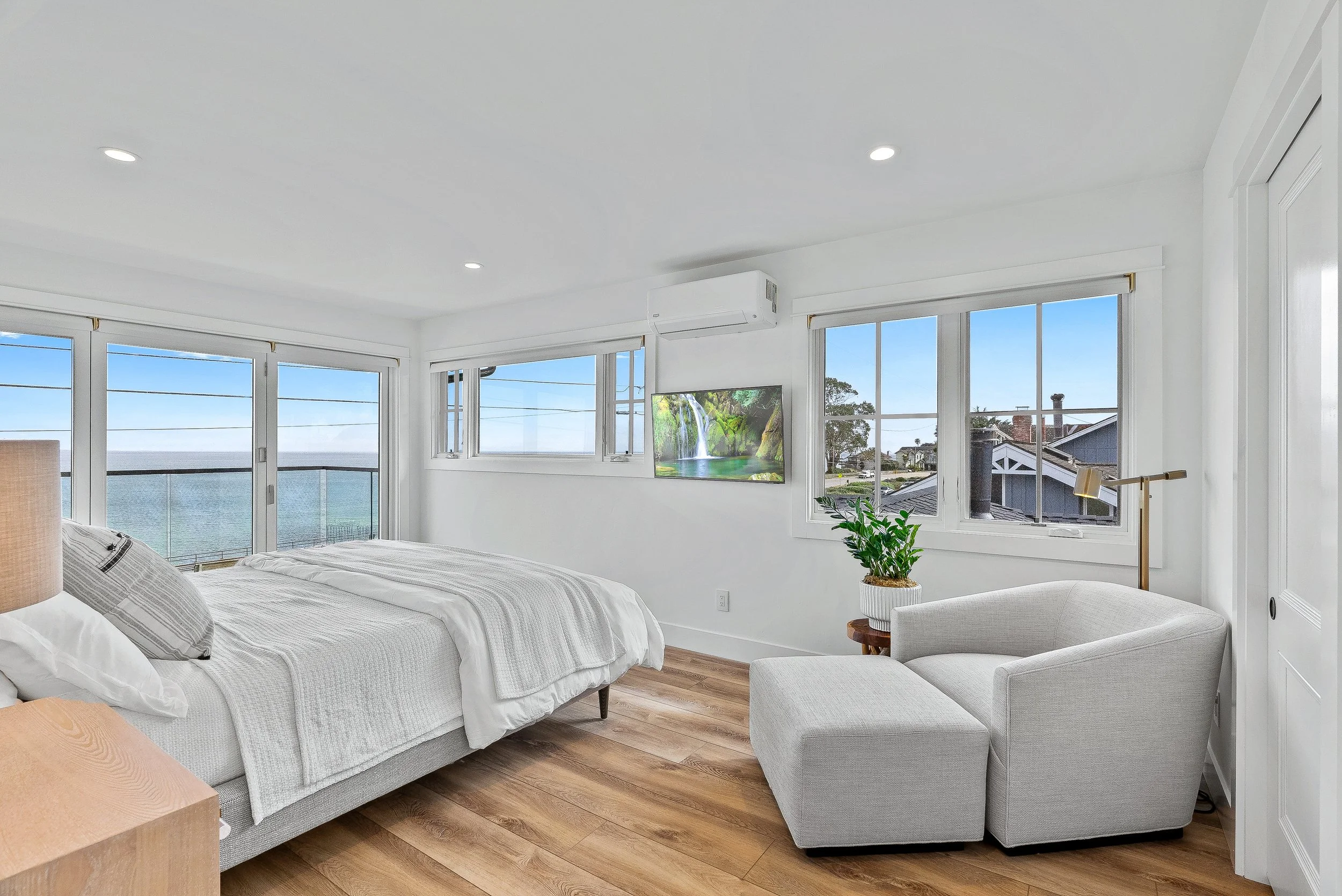 Bright bedroom with large windows showing ocean view, white bedding, a flat-screen TV, a white armchair with an ottoman, a wooden nightstand, potted plant, and wooden floor in Capitola, California.