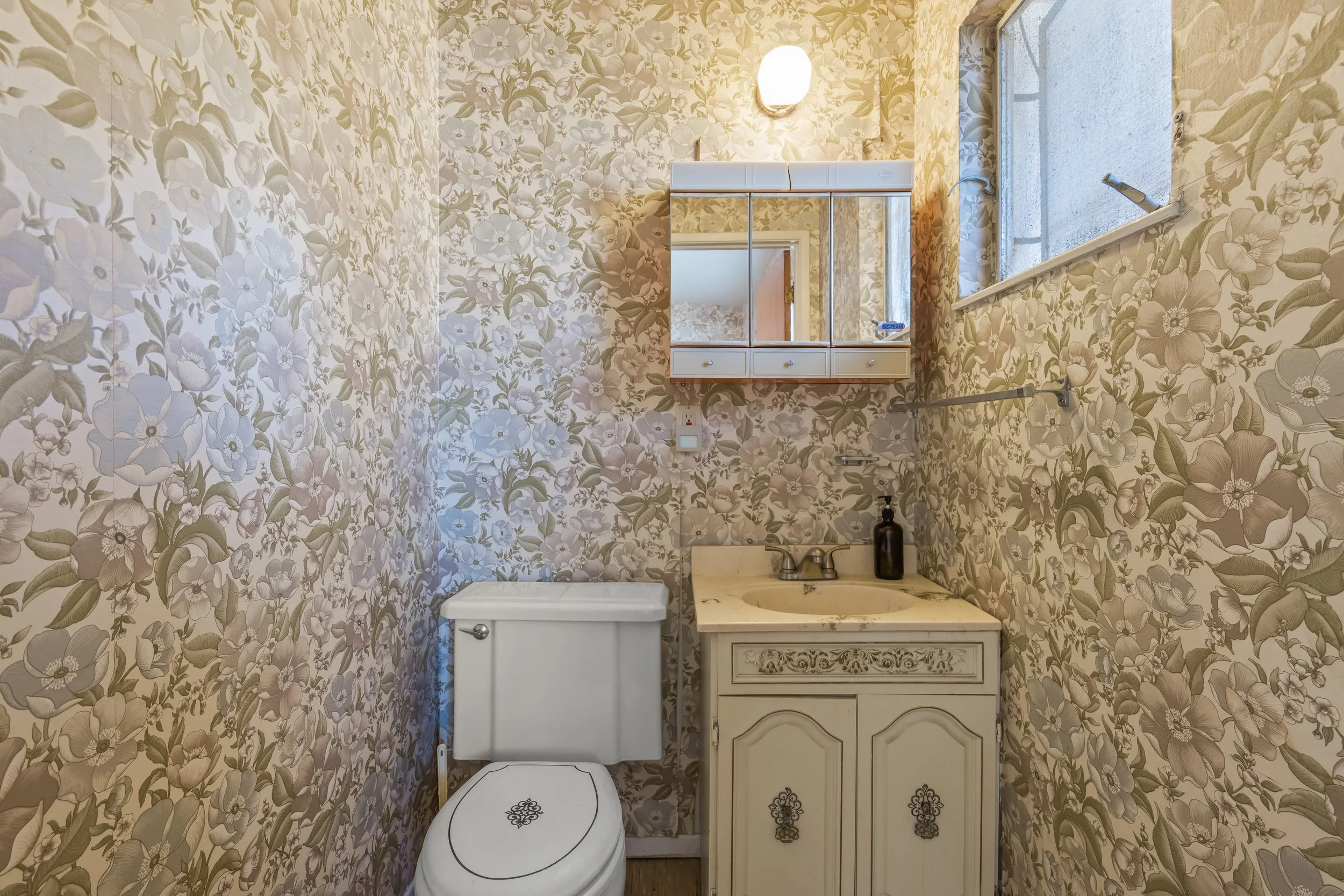 Half bathroom featuring vintage floral wallpaper, single-sink vanity, mirrored medicine cabinet, and natural light from a window. This charming space offers character and potential for updates in a centrally located Santa Cruz County home.