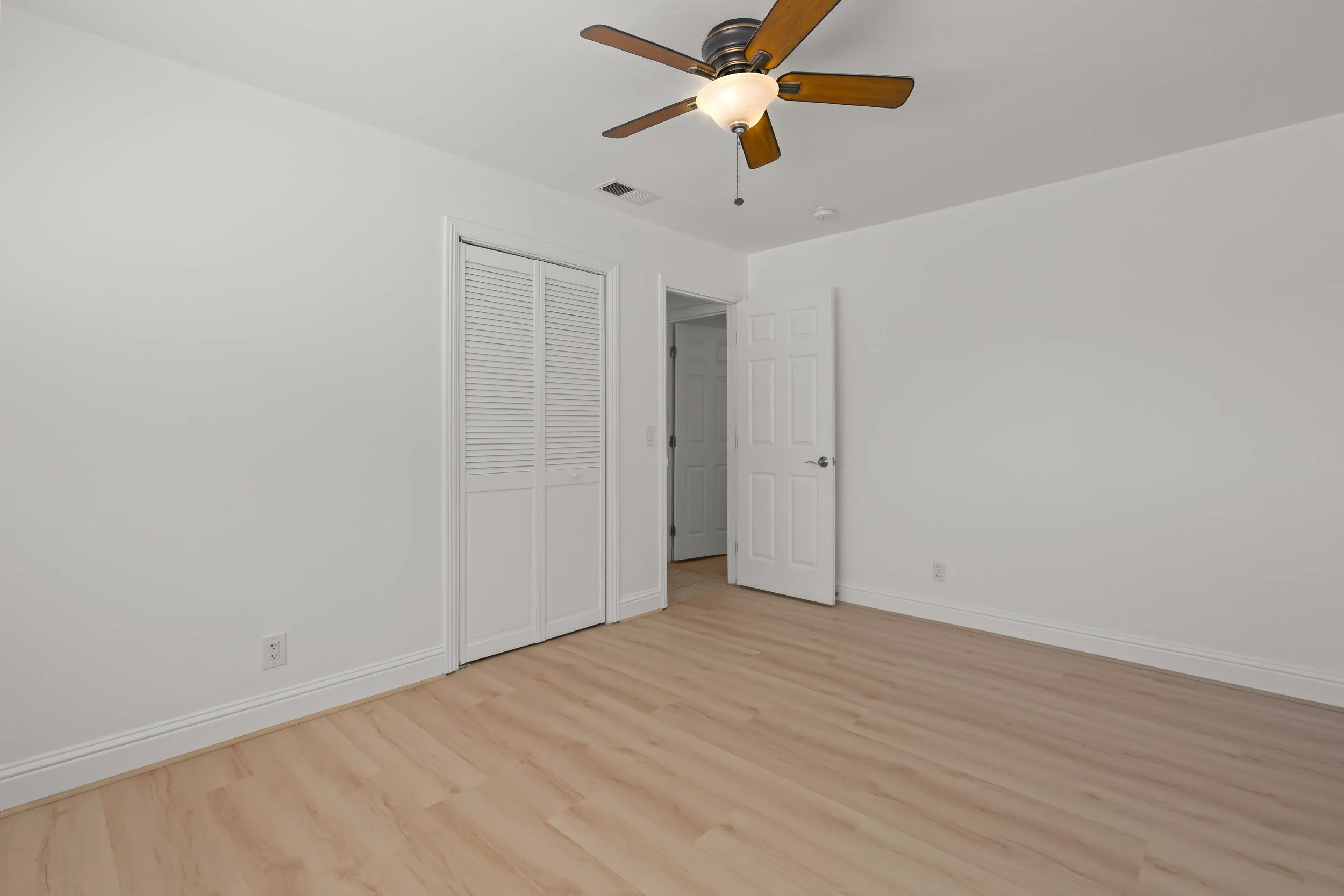Empty bedroom with white walls, wood flooring, a ceiling fan, a closet with louvered doors, and an open door leading to another room.