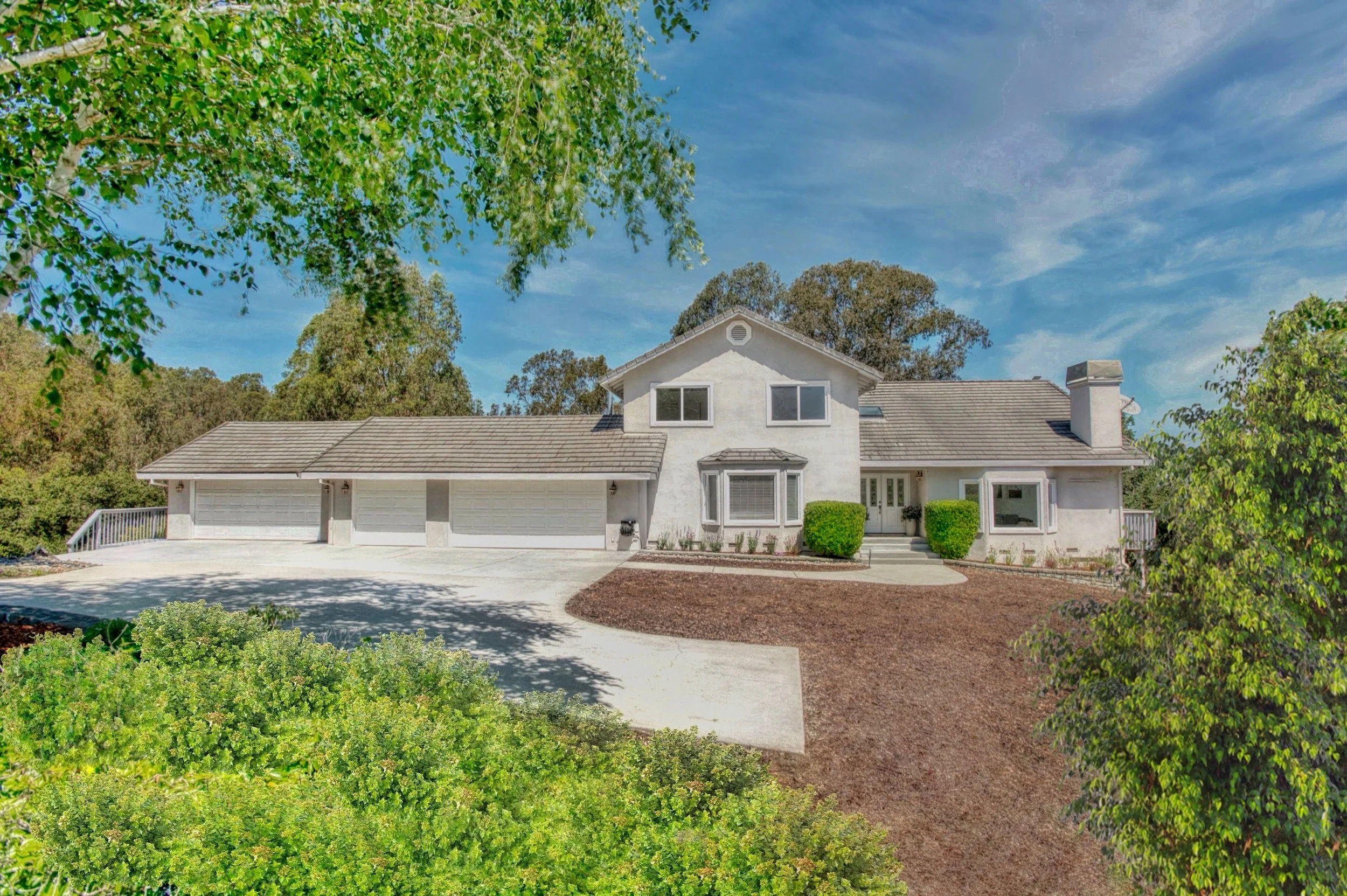 A two-story white house with a three-car garage, surrounded by greenery, shrubs, and trees, under a partly cloudy blue sky.