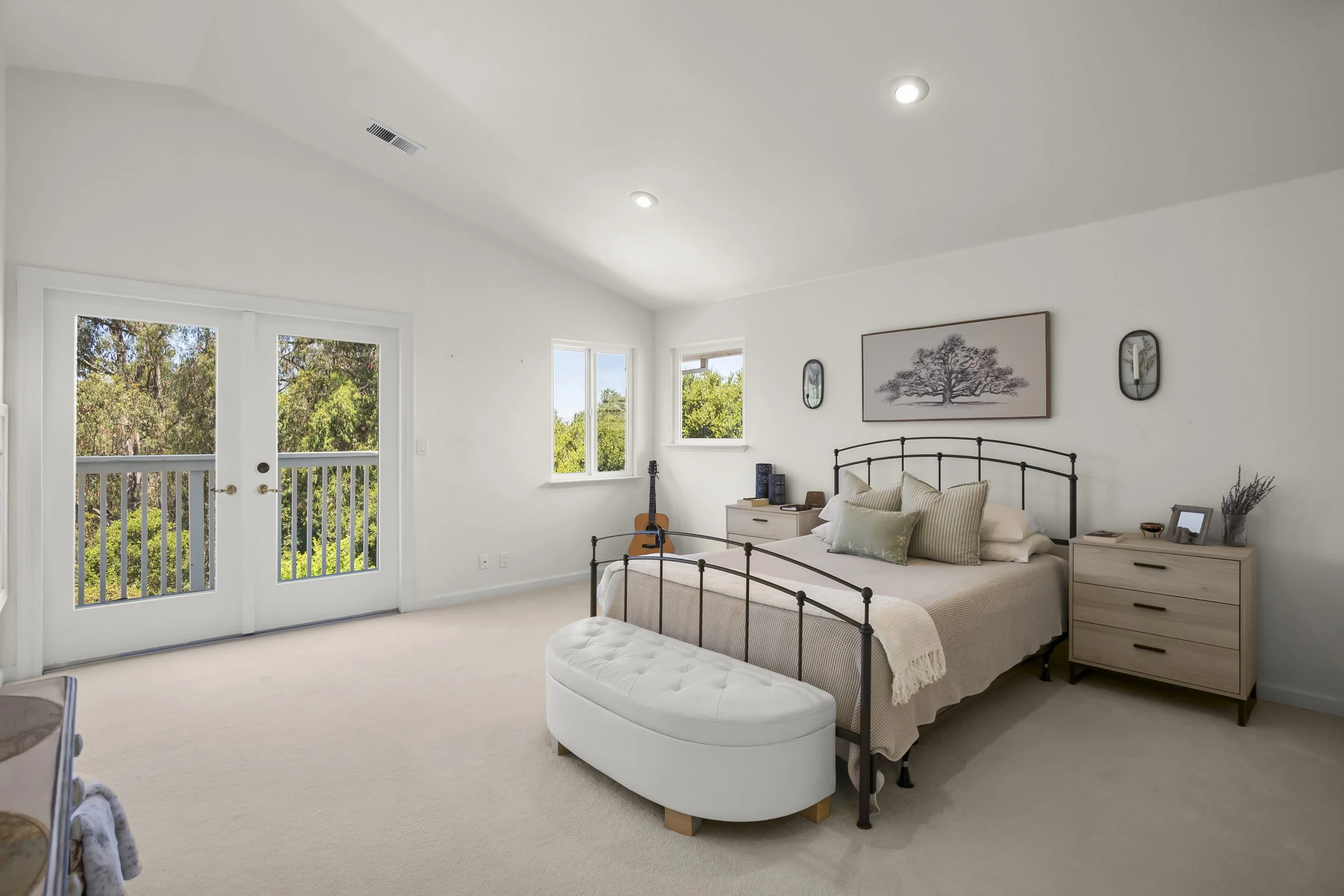 Bright, spacious bedroom with a bed, nightstands, framed artwork, and a guitar, overlooking greenery through windows and a glass door leading to a balcony.