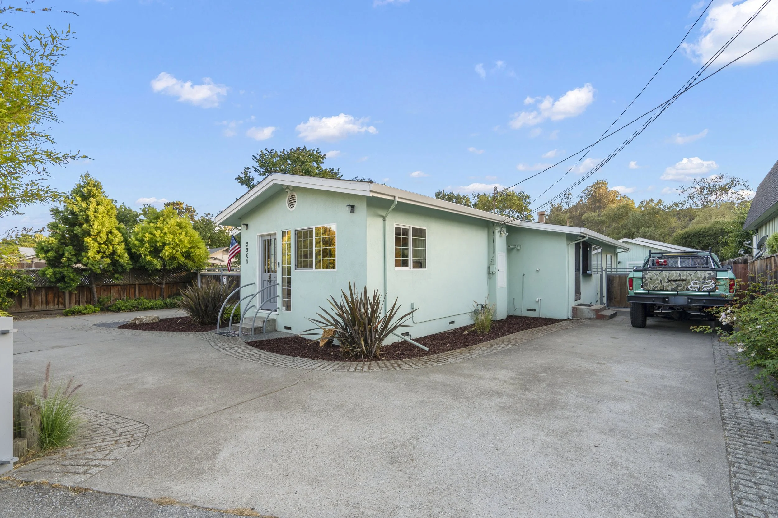 Light green single-story house with small front steps, a driveway, and a pickup truck parked on the right side. There are landscaped plants and a wooden fence in the background, with a partly cloudy sky overhead.