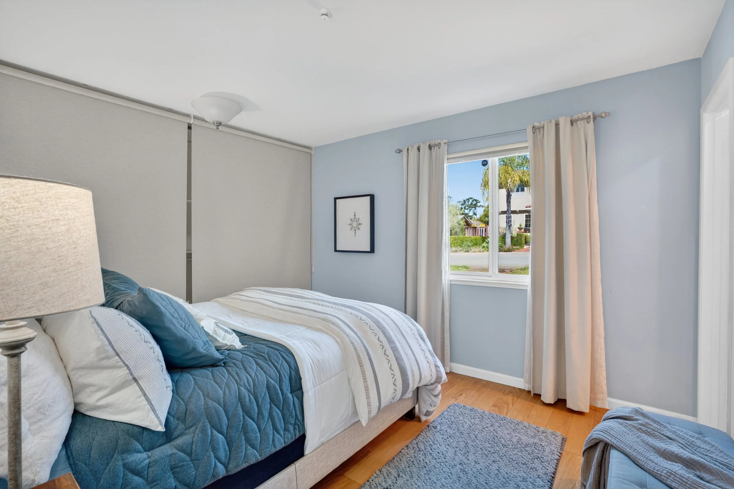 Bedroom with a bed, blue quilt, white and blue pillows, bedside lamp, window with white curtains, and a framed picture on the wall in 1400 Dolphin Dr., Aptos CA.