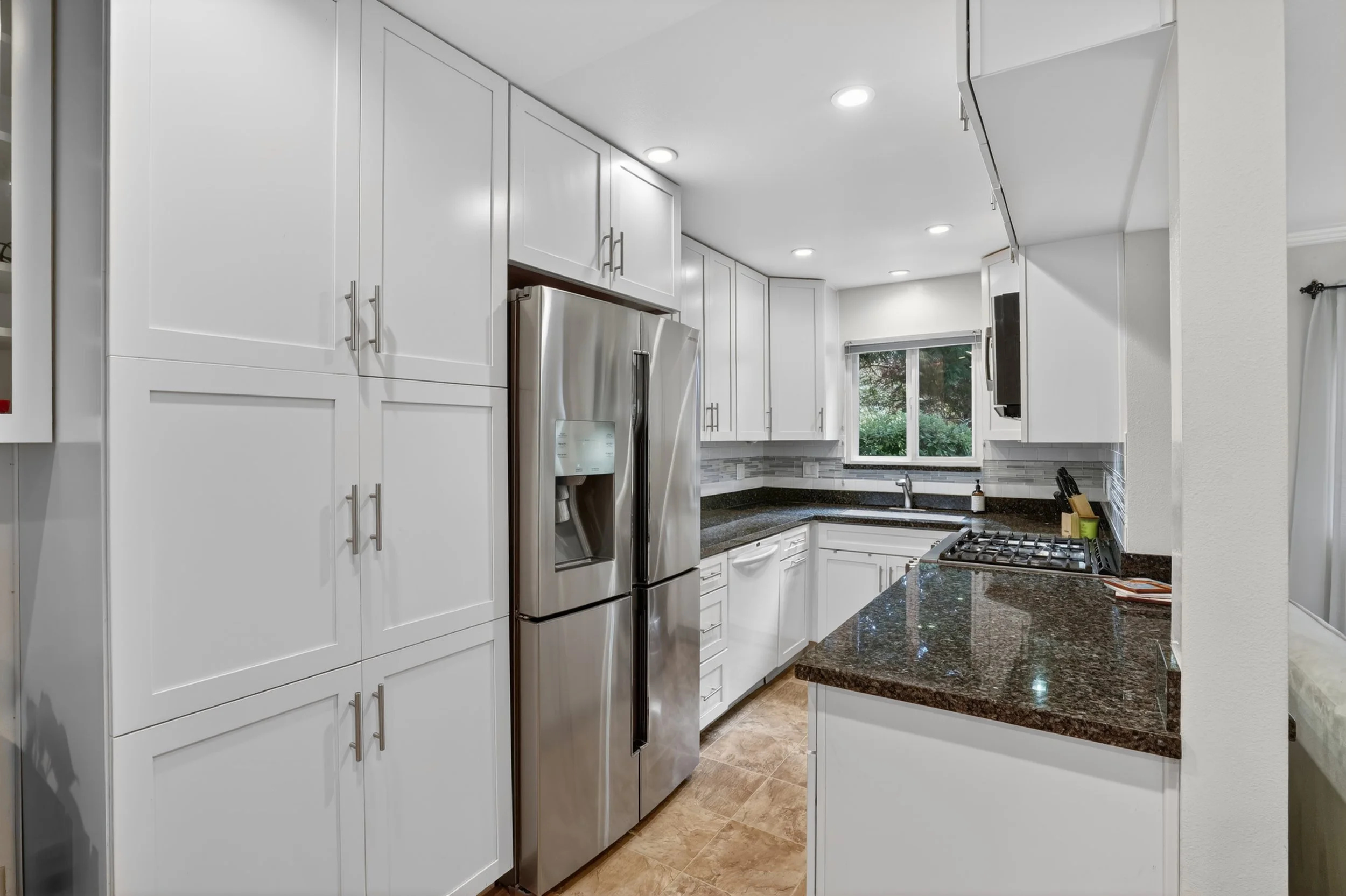 Modern kitchen with white cabinets, black granite countertops, stainless steel refrigerator, gas stove, and window overlooking greenery.