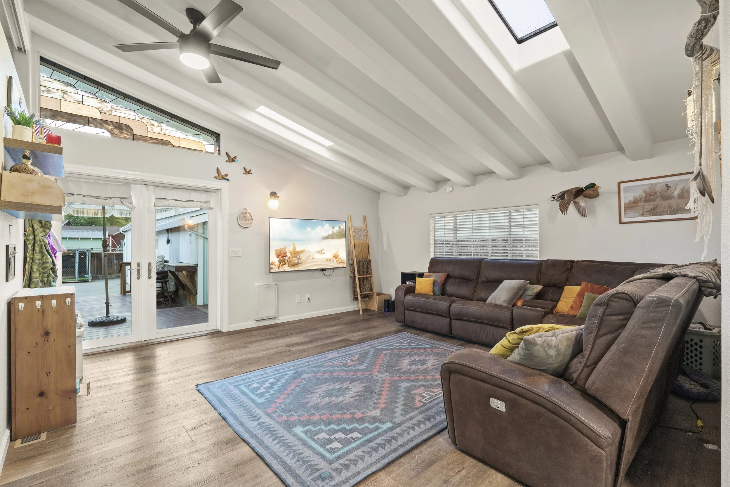 Living room with wood flooring, large sectional sofa with pillows, patterned area rug, wall-mounted flat-screen TV, ceiling fan, skylight, sliding glass door leading to outdoor deck, windows with blinds, and decorative wall hangings including a mount
