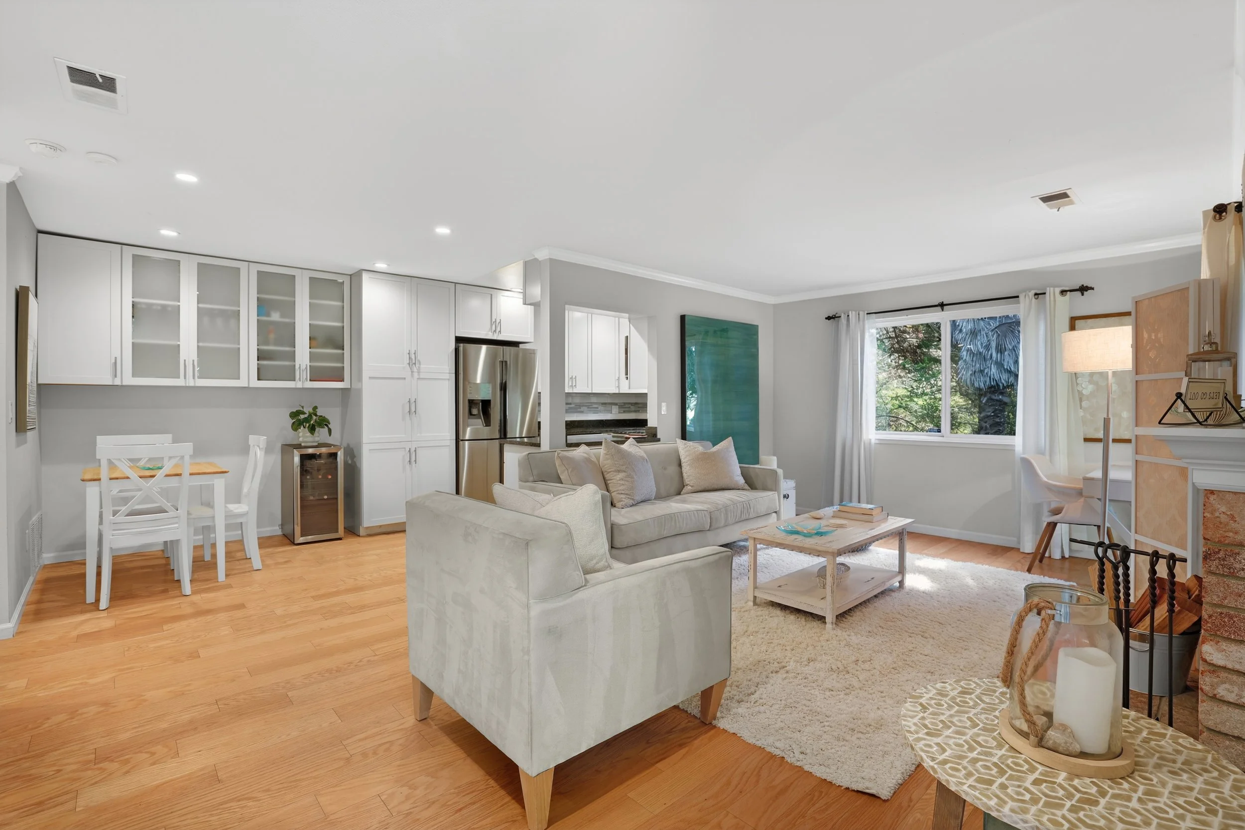 Living room with beige sofa, armchair, coffee table, and area rug; kitchen with white cabinets and stainless steel refrigerator; large window with curtains showing trees outside; decorative items and a small desk nearby.