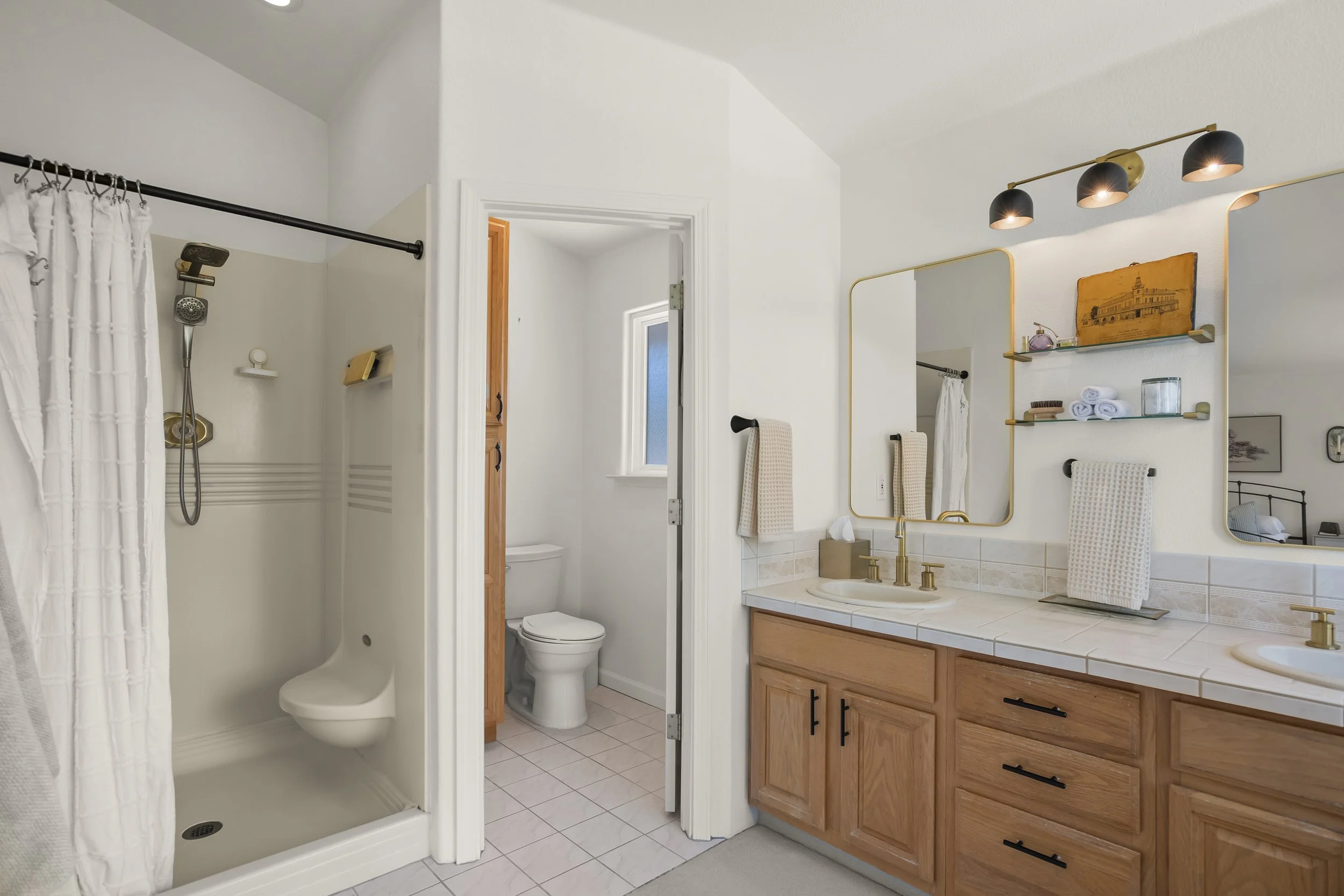 Bathroom with double vanity, large mirror, black light fixtures, wooden cabinets, and a separate toilet and shower area.