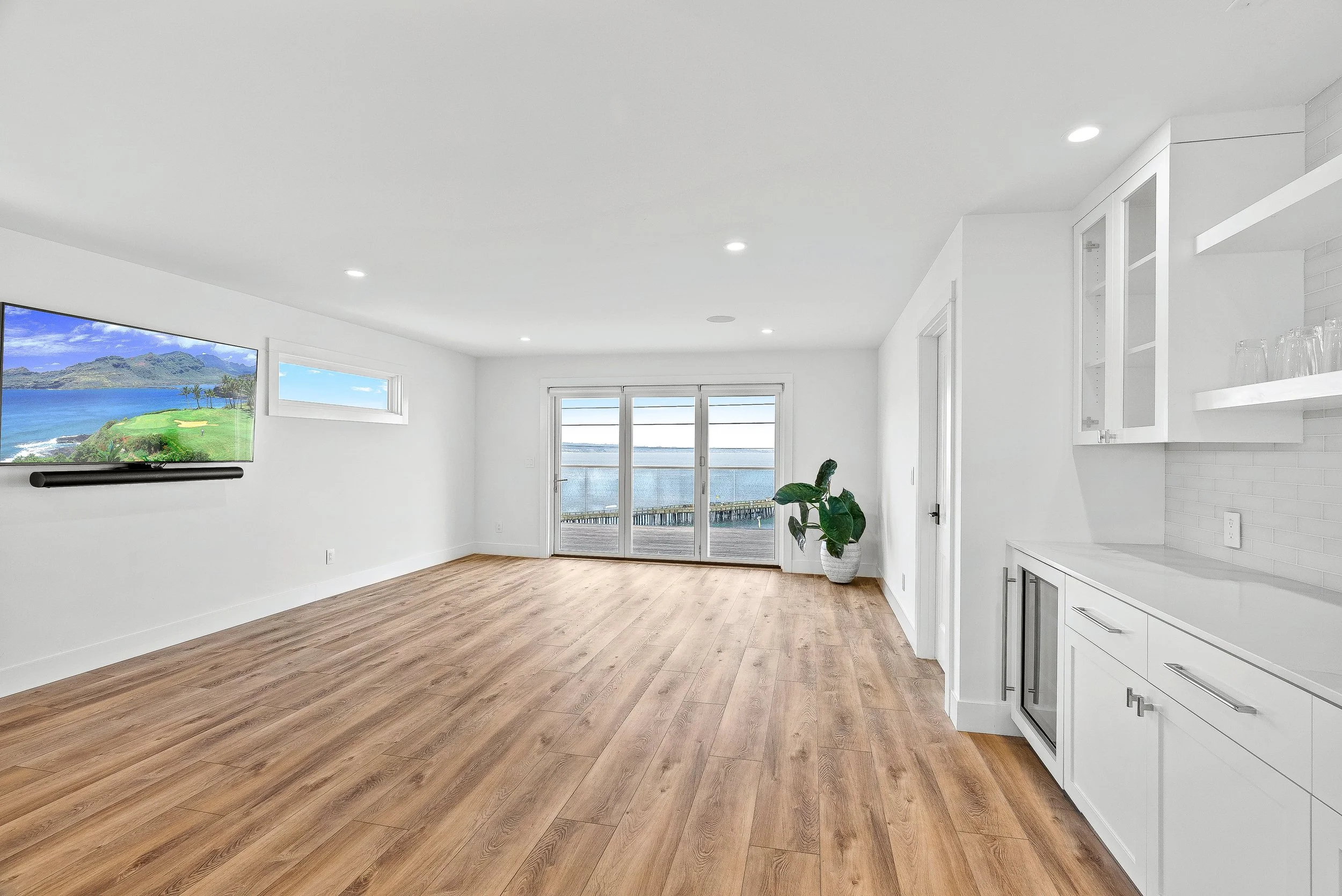 Bright living room with white walls, hardwood flooring, sliding glass doors to a balcony with an ocean view, and a wall-mounted TV showing a scenic landscape in Capitola, California.