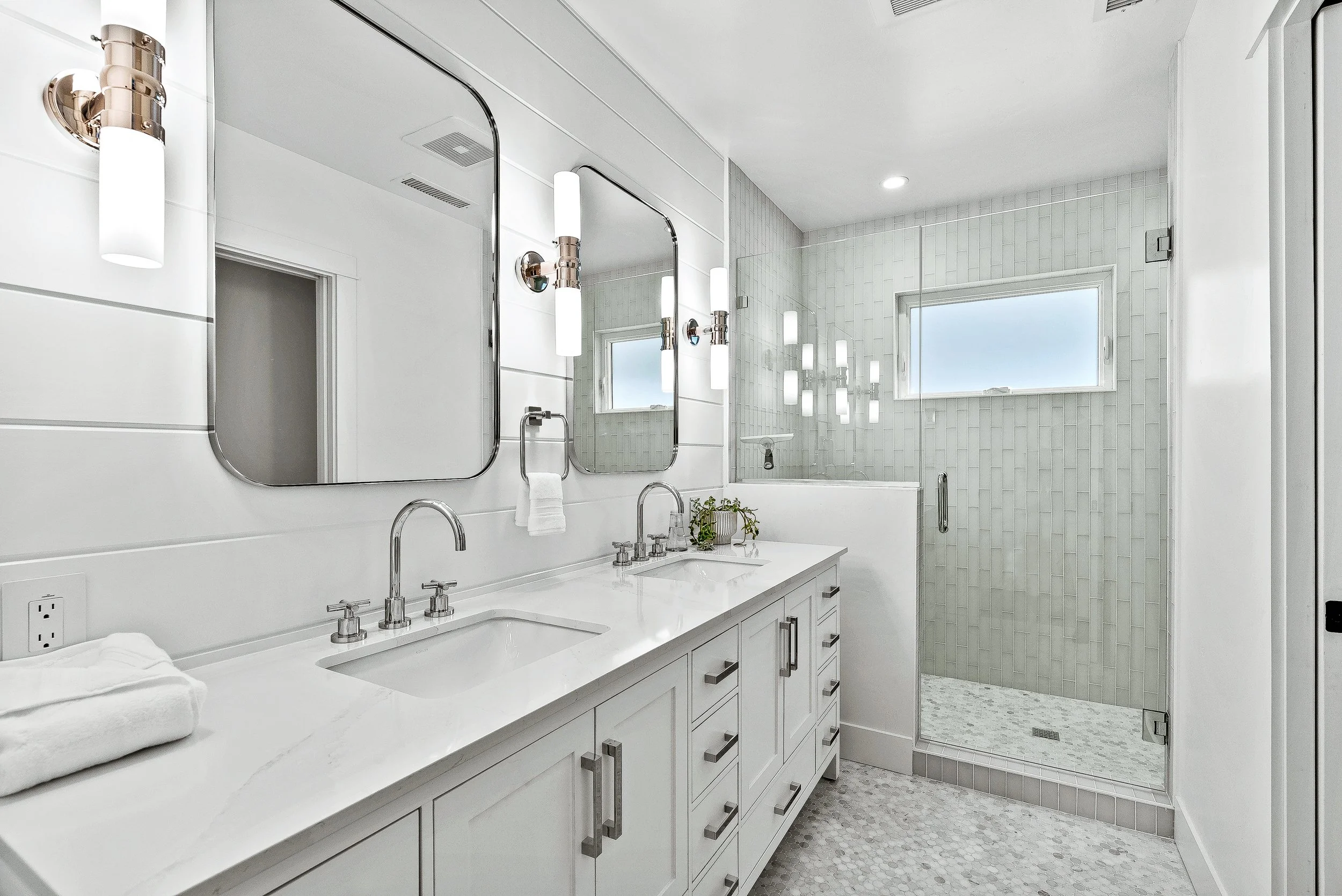 Modern white bathroom with double vanity, large mirrors, wall-mounted lights, a glass-enclosed shower with a window, and a patterned tile floor in Capitola, California.