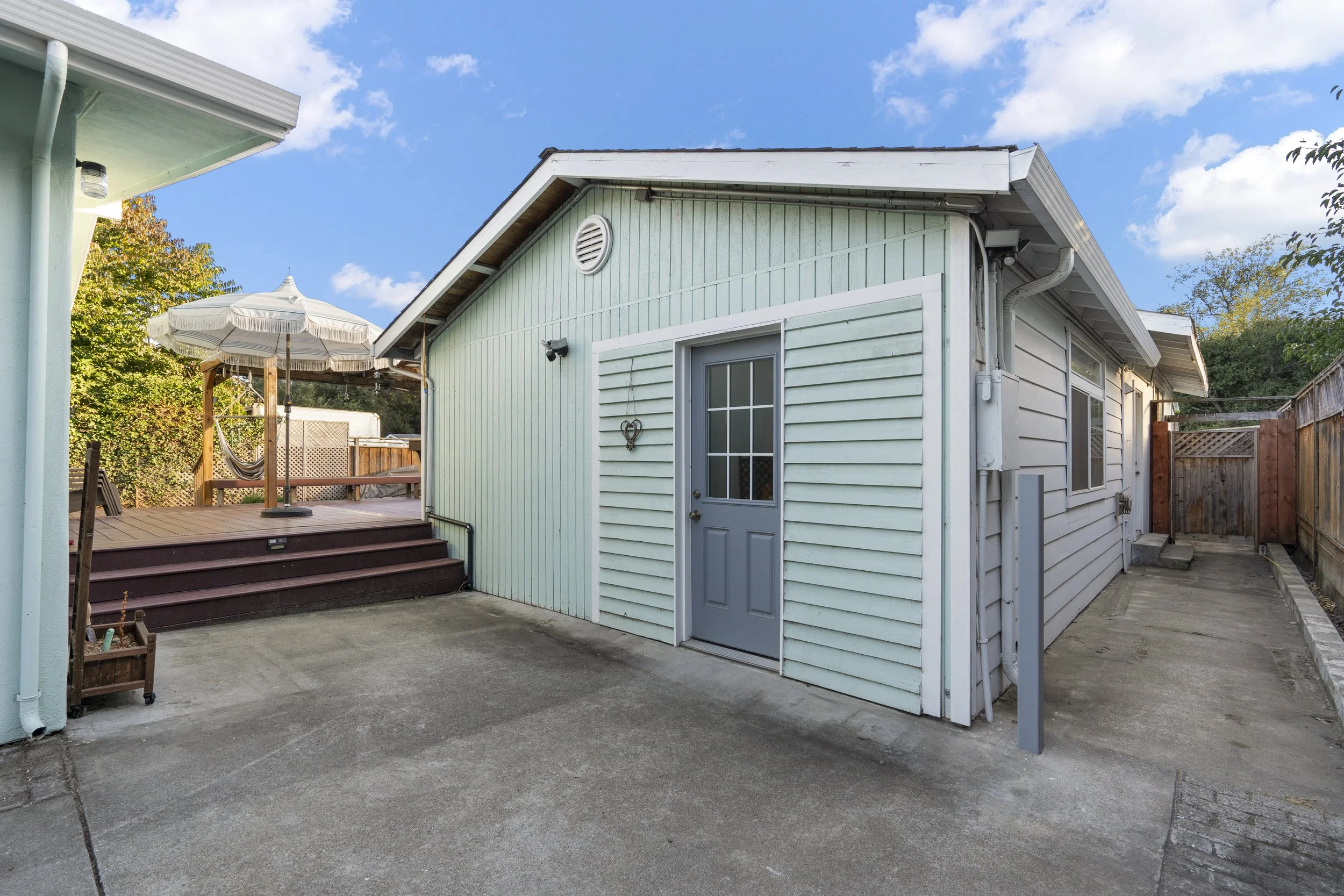 Backyard with a small house, wooden deck with an umbrella and hammock, concrete patio, and fenced yard under a partly cloudy sky.