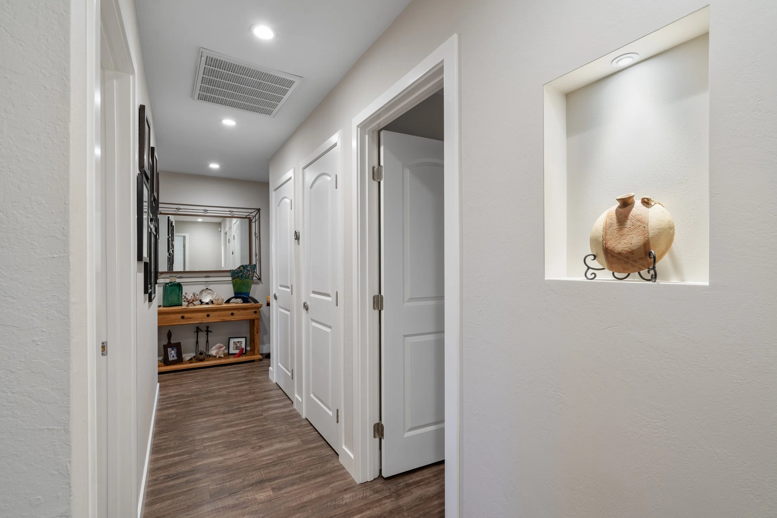 Hallway with white walls, wooden floor, white doors, a small wall niche with a decorative vase, and a mirror reflecting a console table with decorative items at the end of the hall.