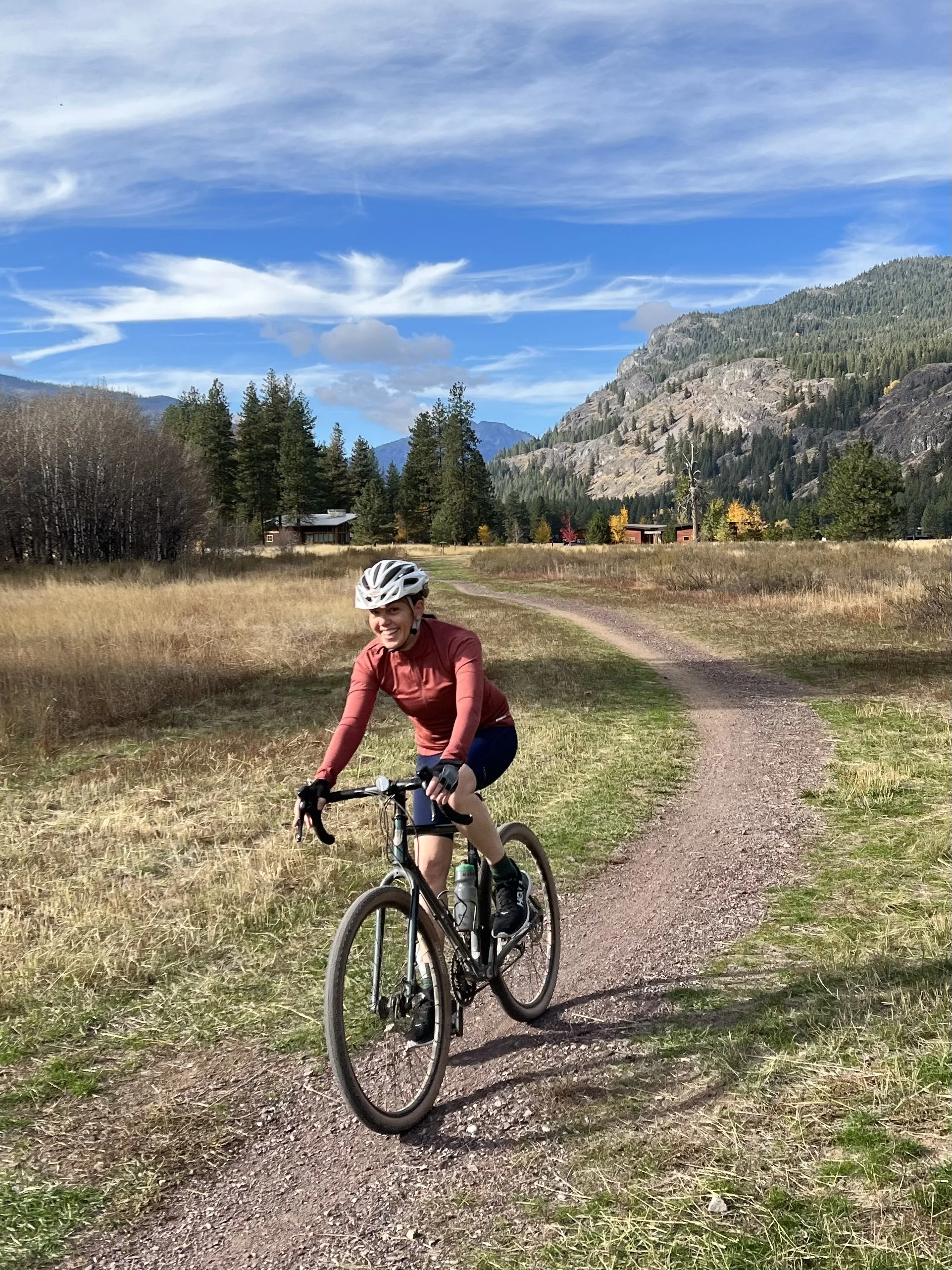 Seattle based leadership coach and consultant, Meredith Lohr, is cycling on a dirt trail in a scenic mountainous area with trees, grass, houses, and a partly cloudy sky.