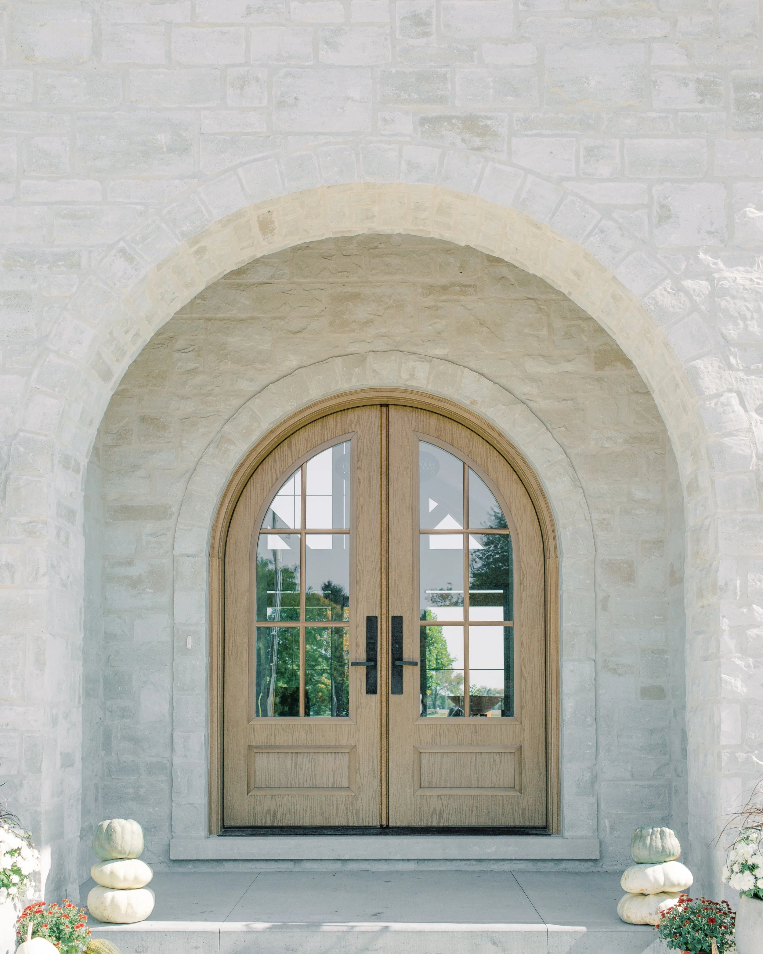 Wooden double doors with glass panes, set in stone wall with an arched entrance, and decorated with pumpkins and flower pots at the porch.