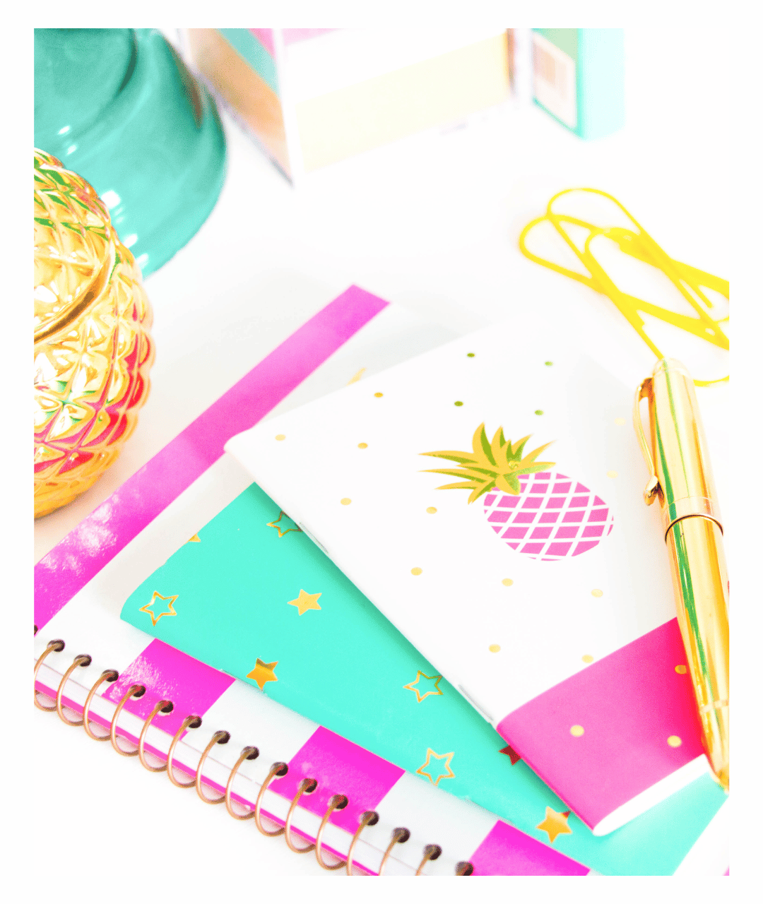 A colorful desk with notebooks, a gold pen, a yellow paper clip, a gold pineapple-shaped container, and pastel-colored storage boxes.