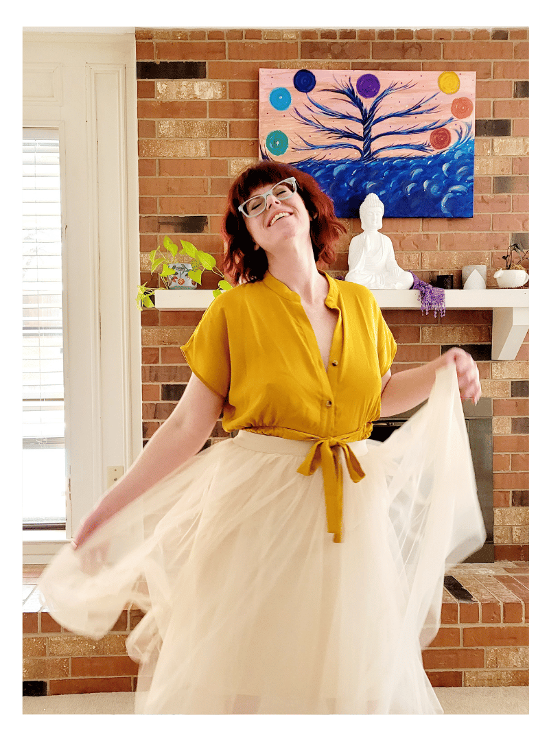 Kate Smith wearing a yellow top and a cream-colored tulle skirt smiling and holding the skirt out in a bright living room with a brick fireplace, a brick wall, a white shelf with plants and decor, and a colorful tree painting in the background.