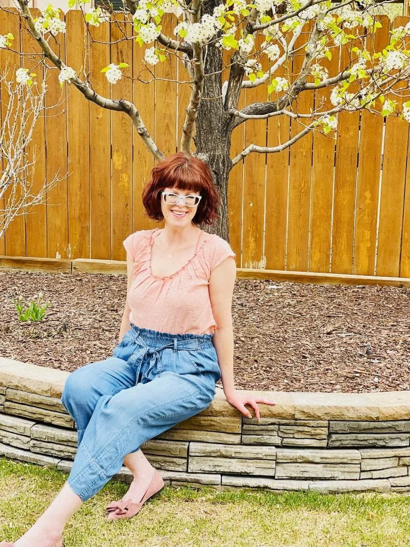 Website Designer - Kate Smith pictured in a pink top and jeans sitting in front of a flowering tree