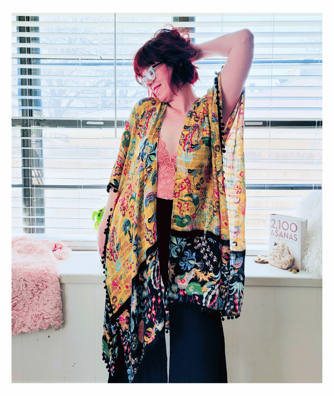 Kate Smith, Website Designer is standing in front of a large window with blinds, wearing glasses, colorful floral and patterned kimono, pink lace top, and black pants, smiling with one arm raised and her hand behind her head.
