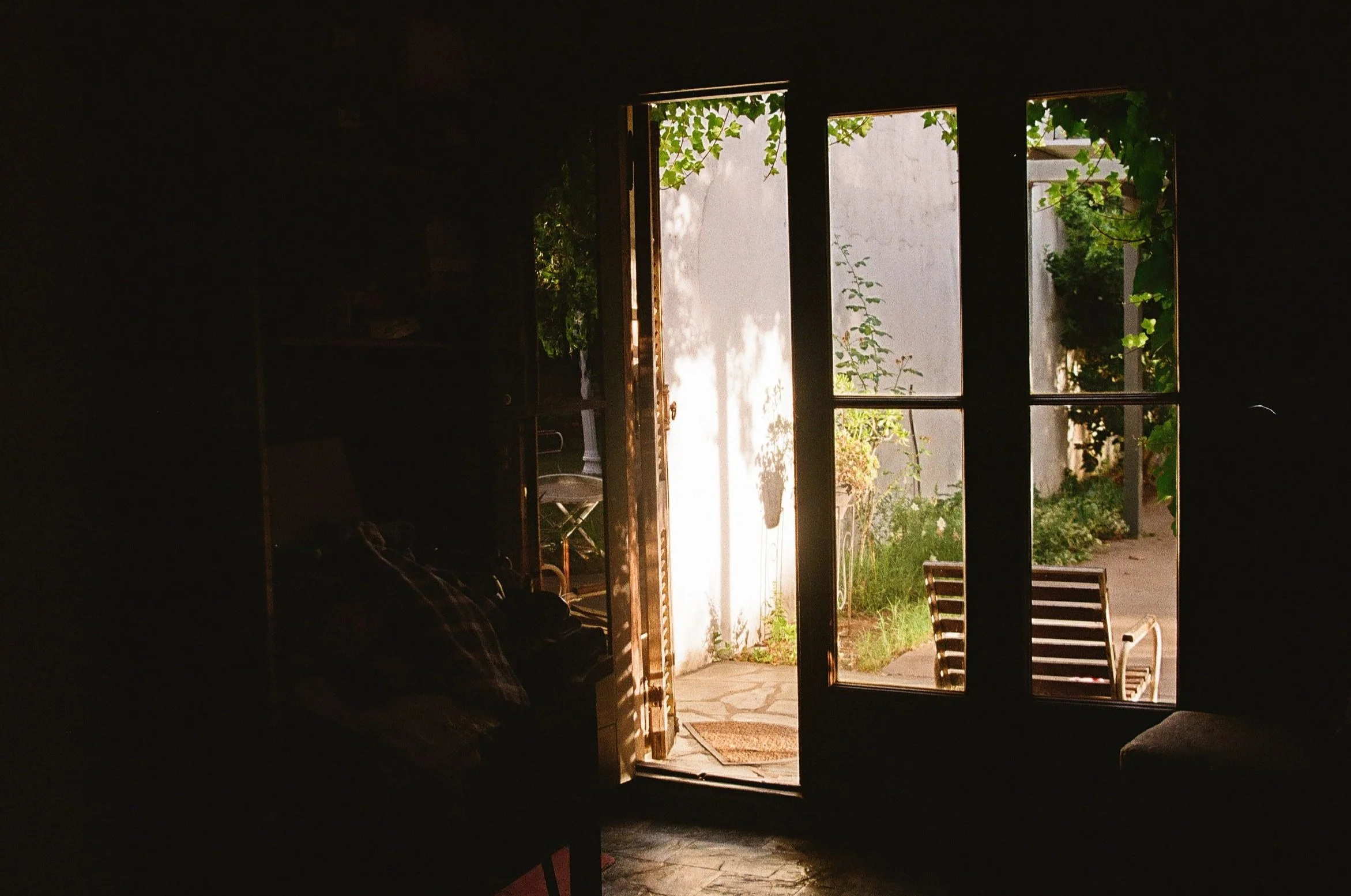 Open door leading to a sunlit outdoor patio with plants, chairs, and a white wall.