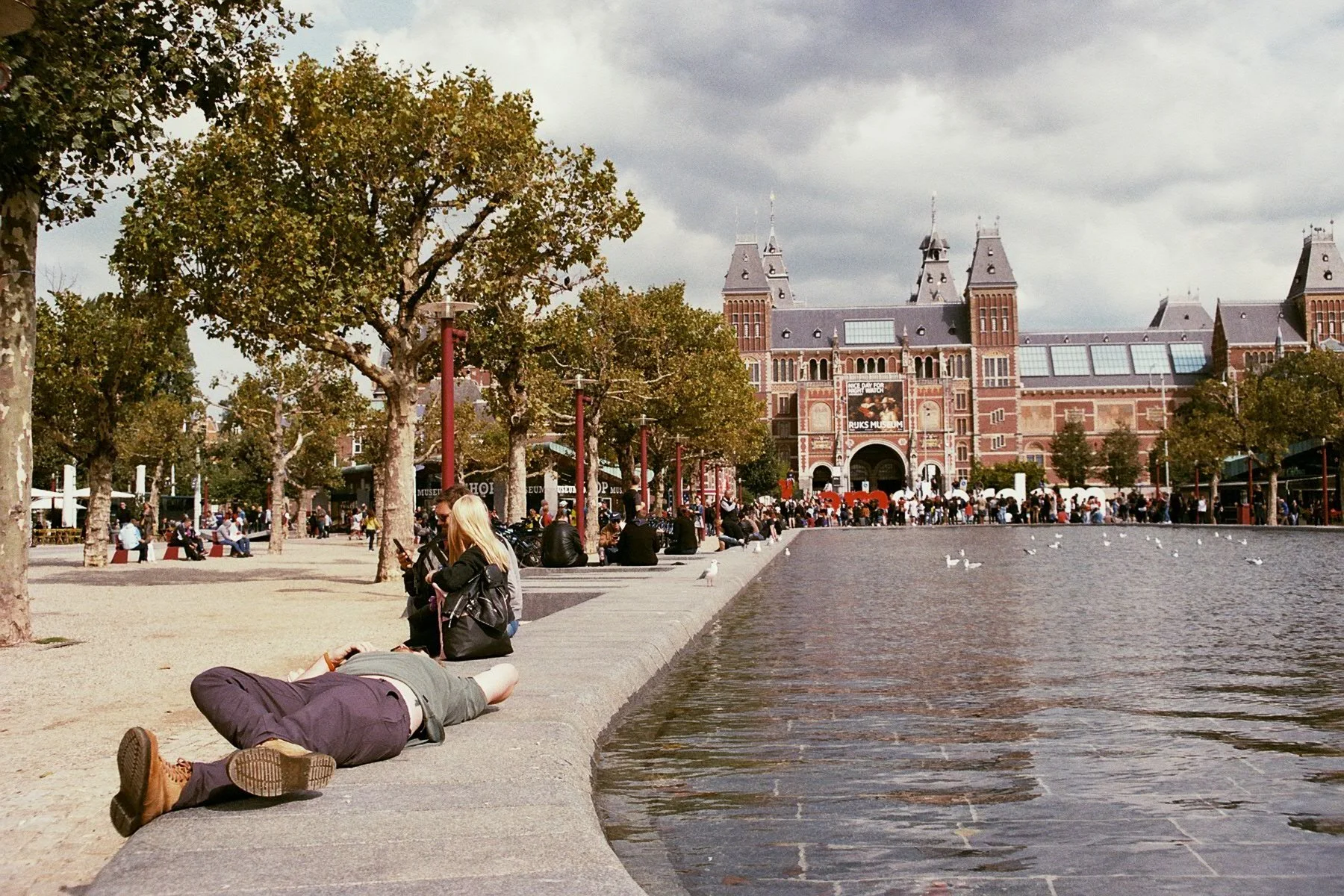 People sitting and walking along a waterfront promenade with a large historic building resembling a castle or museum in the background, trees lining the walkway, and seagulls near the water under cloudy skies.