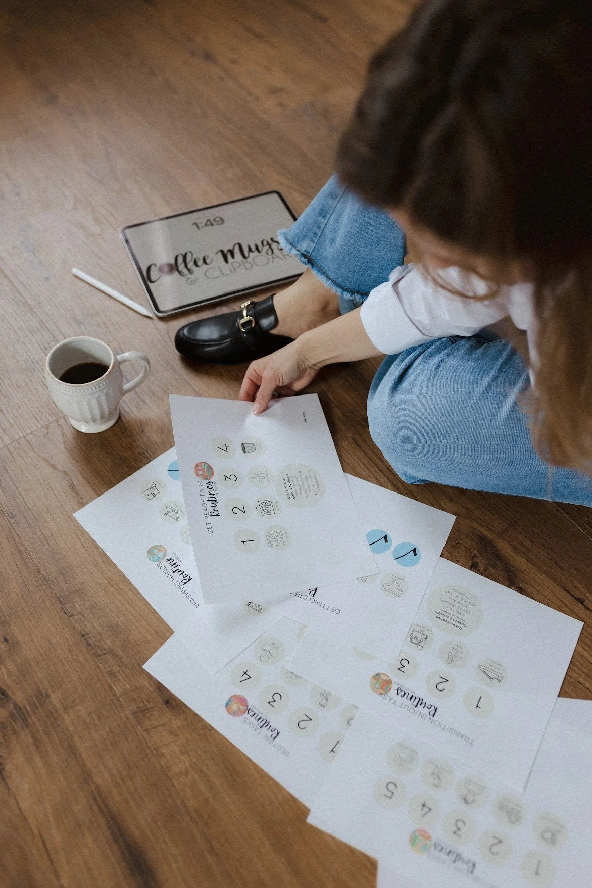 Person sitting on a wooden floor with a cup of coffee, holding a sheet of paper with numbered and illustrated tasks. Several other sheets with similar content are spread on the floor. A digital tablet and a stylus are nearby, with the tablet displaying the time 1:49 and text that reads 'Coffee Mug & Cup'.