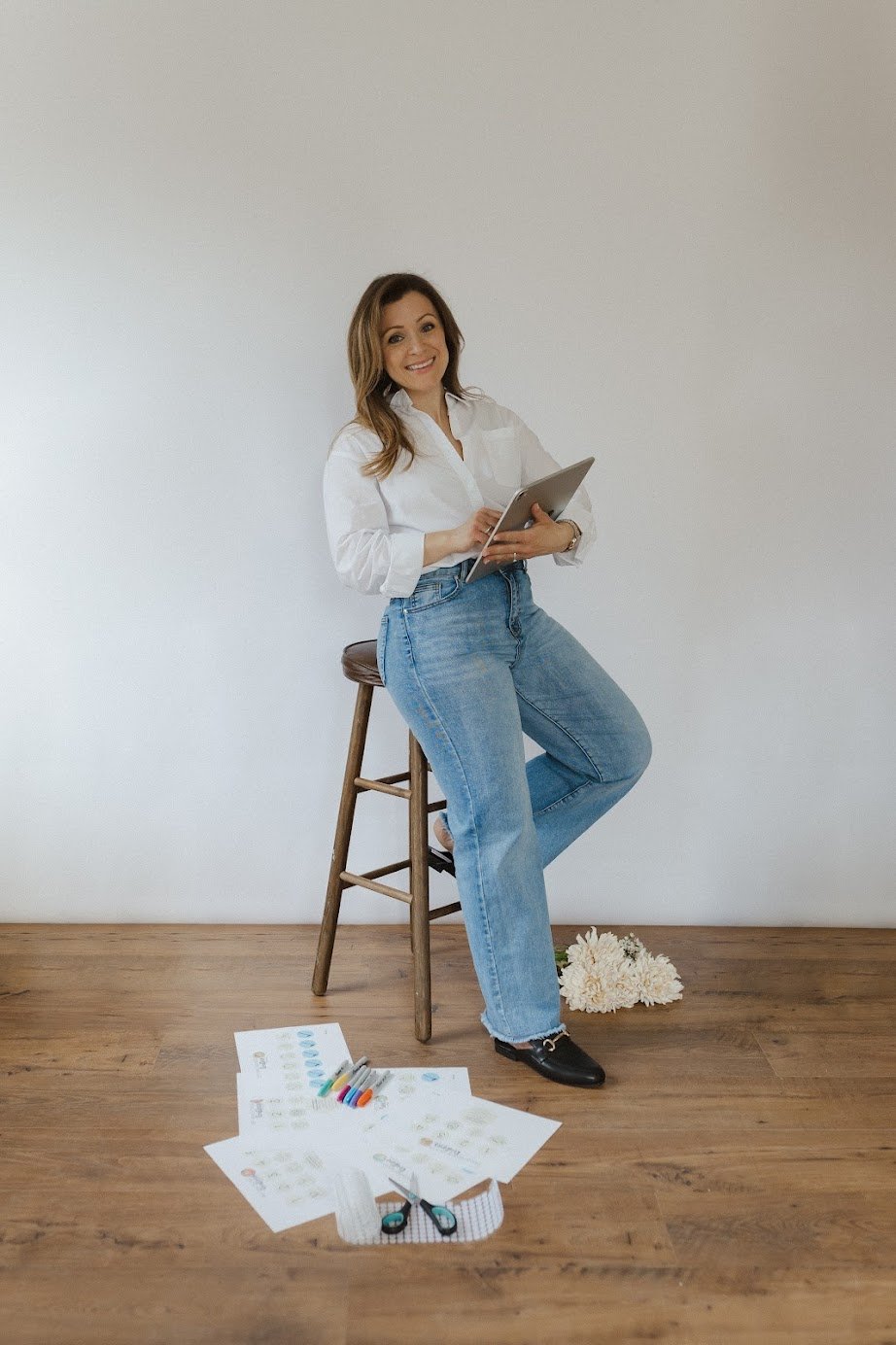 Woman sitting on a stool with a tablet, surrounded by papers, markers, scissors, and a tape measure on a wooden floor, against a plain white wall.