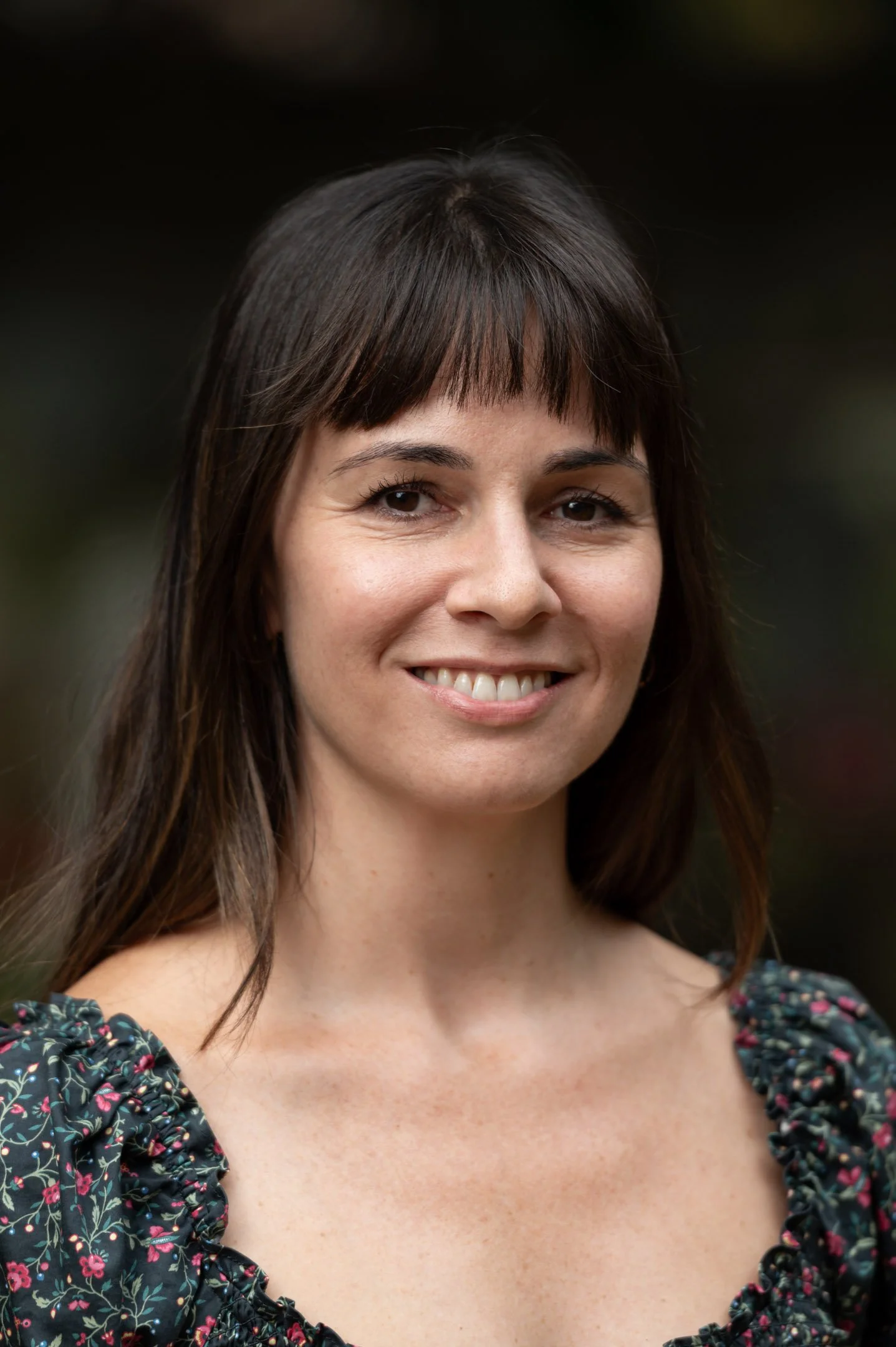 A woman with long brown hair wearing a floral blouse and pearl earrings, smiling outdoors with green foliage in the background.