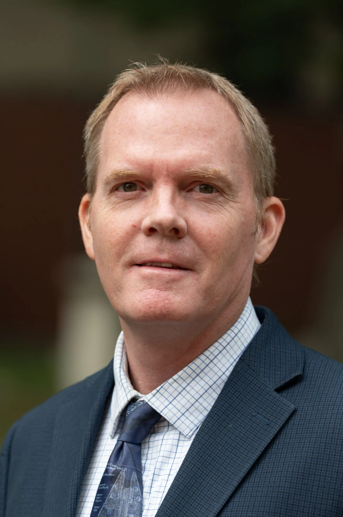 A man with short light brown hair wearing a navy blue polo shirt, standing outdoors with green foliage and a stone wall in the background.
