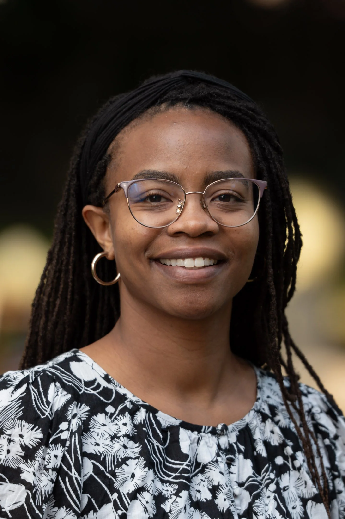 Person smiling outdoors with greenery in the background.