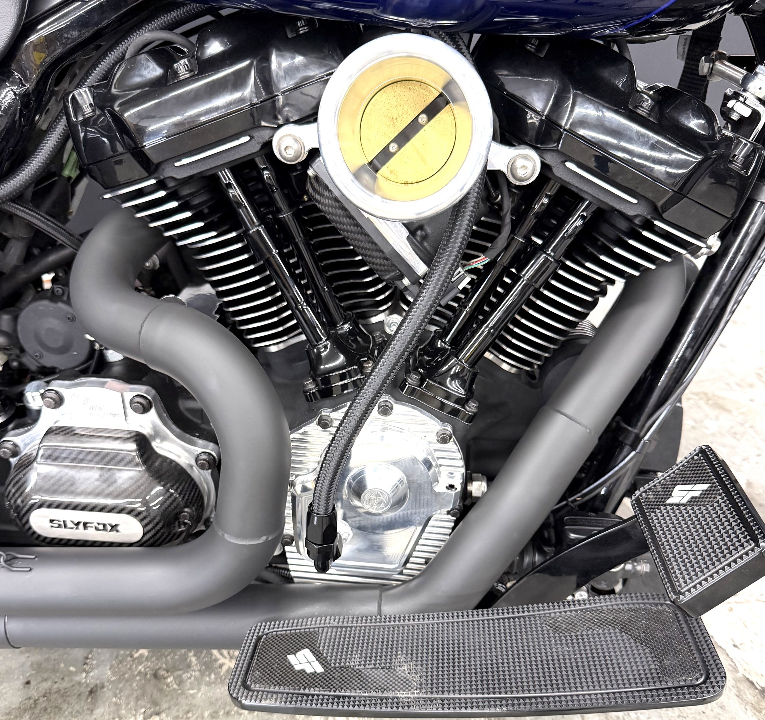 Close-up of a motorcycle engine with various metal and carbon fiber parts, including hoses, bolts, and a yellow circular component at the center.