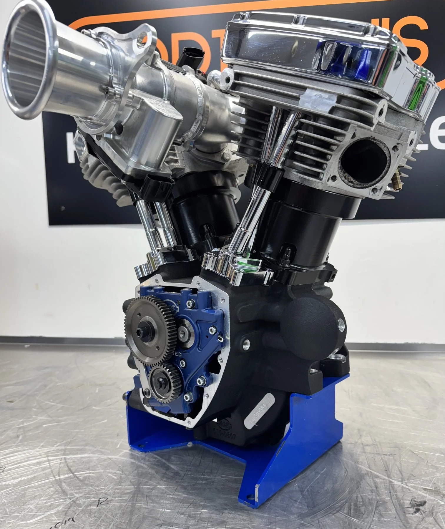 A detailed close-up of a high-performance internal combustion engine with chrome and black components displayed on a blue stand, set against a black background with orange and white partially visible text.
