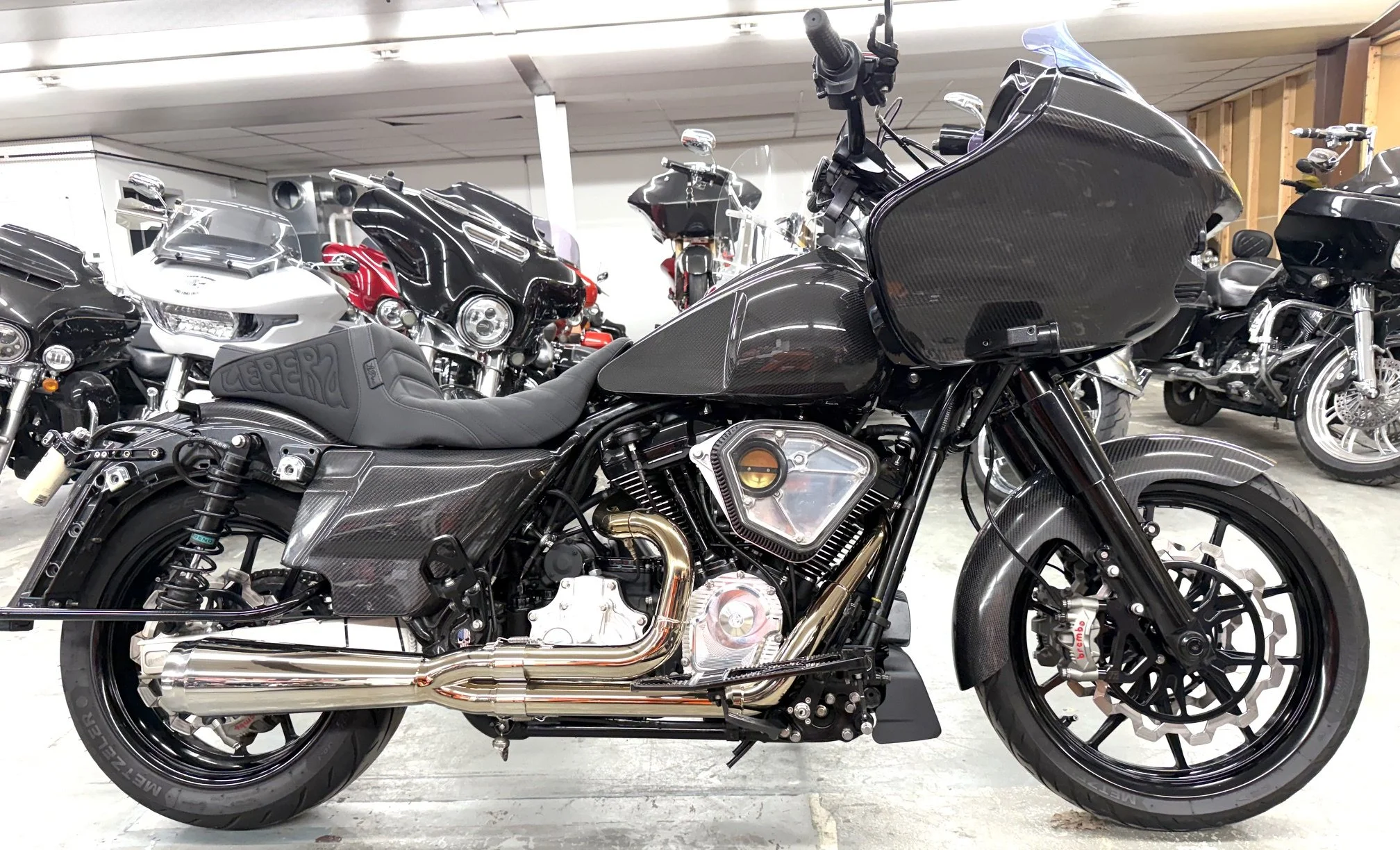 A collection of motorcycles parked indoors, with a black motorcycle in the foreground, featuring a carbon fiber body, chrome exhaust, and black wheels, surrounded by other motorcycles in various colors including white, red, and black.