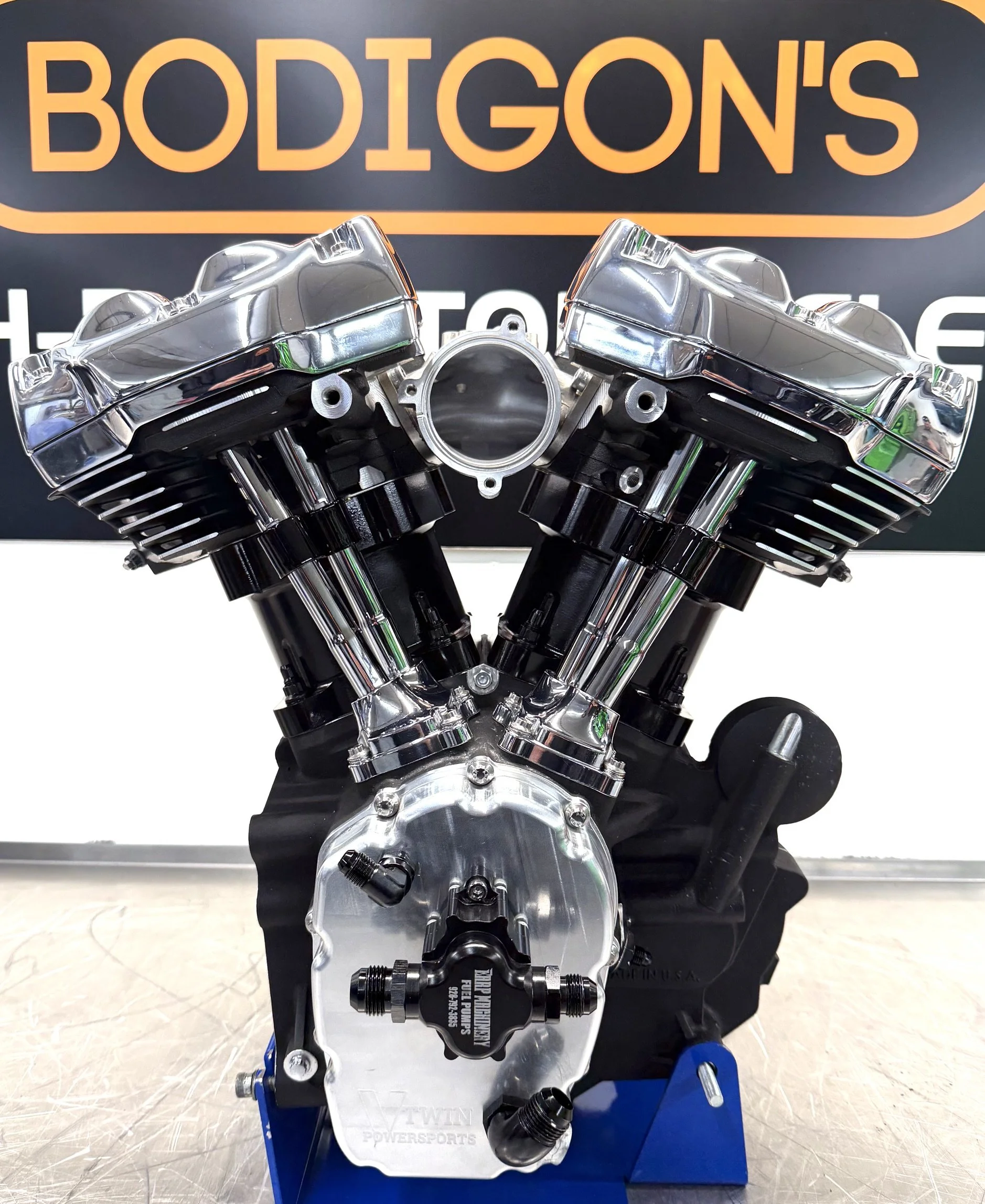 Close-up of a high-performance engine part, likely a cylinder head, mounted on a display stand. The background features a sign with the text 'BODIGON'S' in large orange letters.